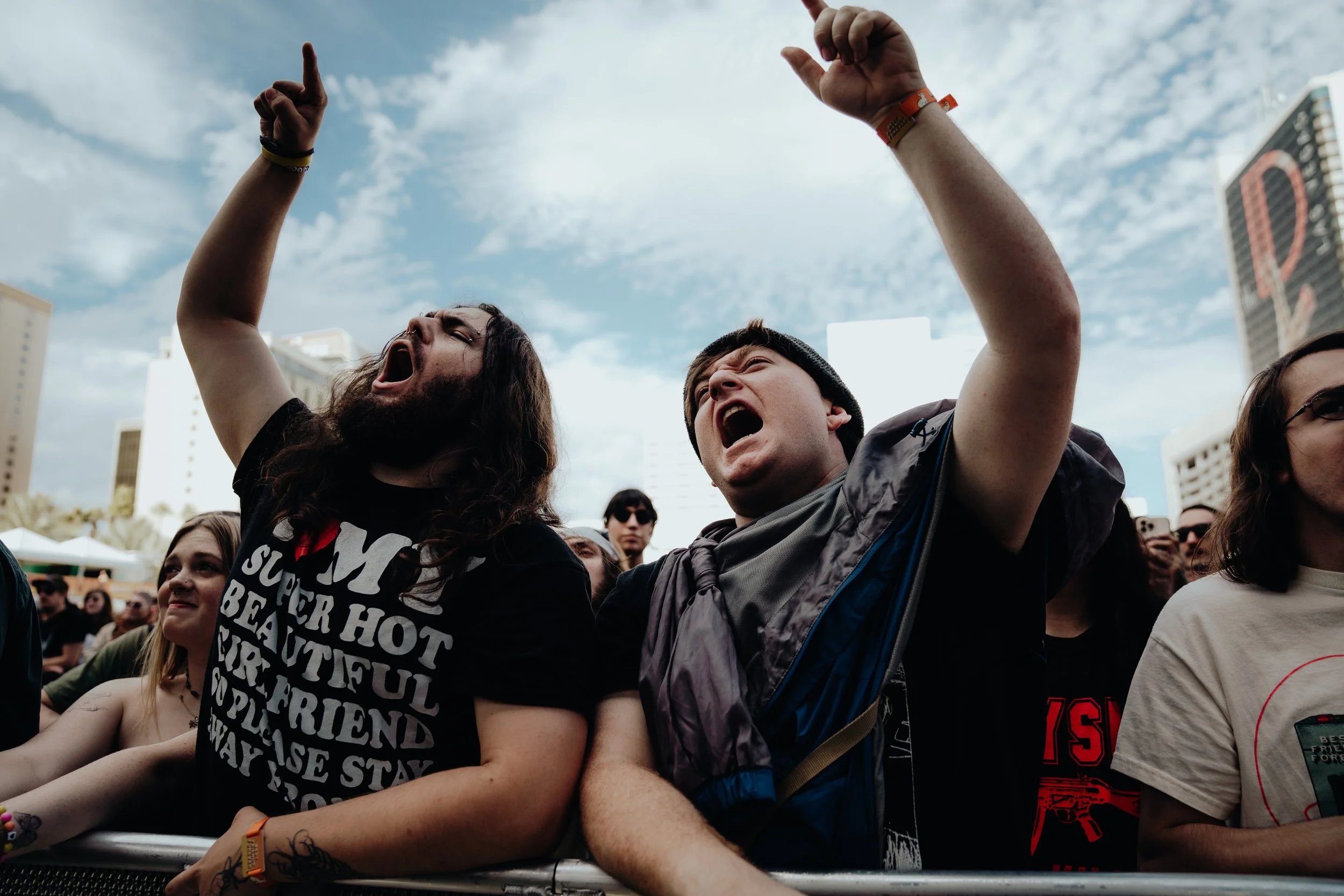 People at a concert or outdoor event, showing excitement and enthusiasm, with some raising their hands and cheering, under a partly cloudy sky.