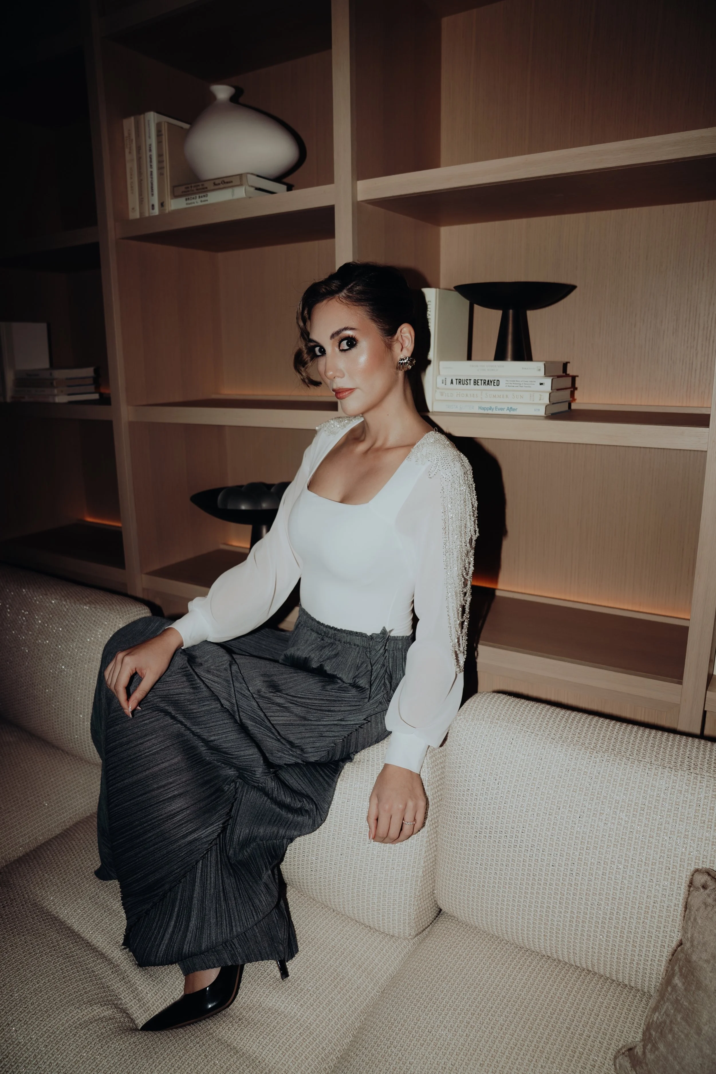 A woman dressed in a white top and gray skirt sitting on a beige sofa in front of a wooden bookshelf.