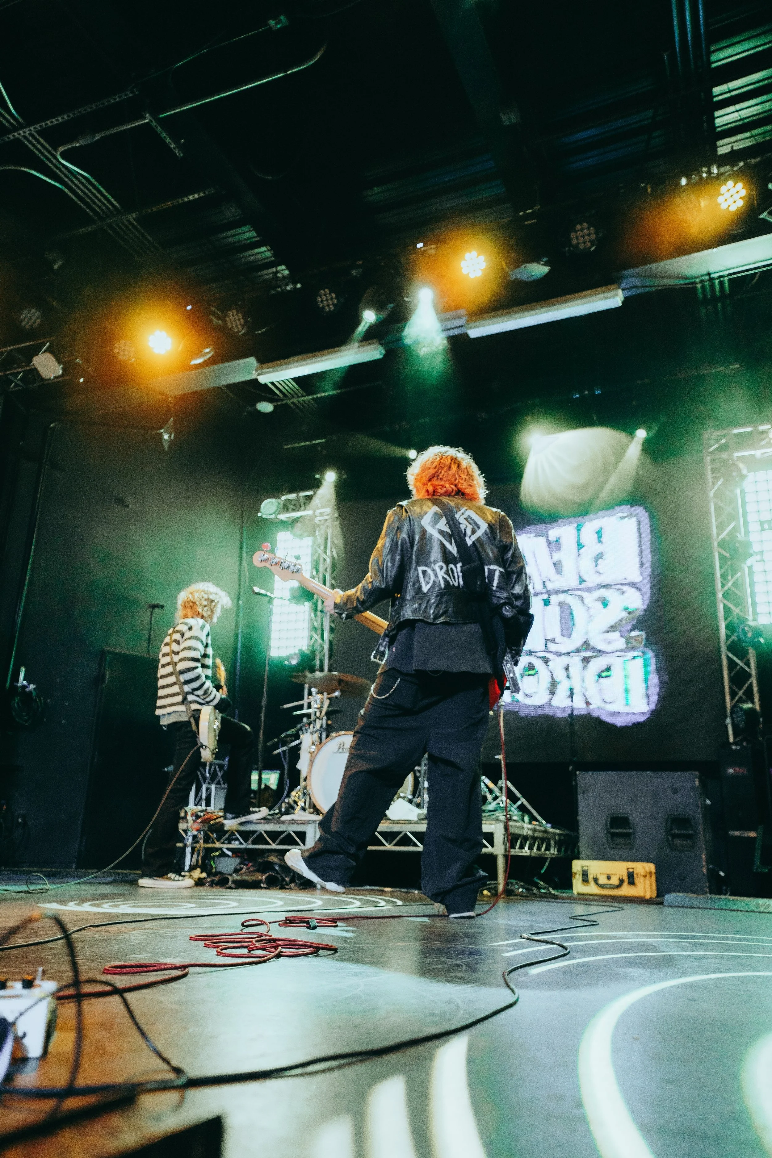 Band performing on stage in a music venue with colorful lighting, including yellow and green hues, and a large screen in the background displaying text.