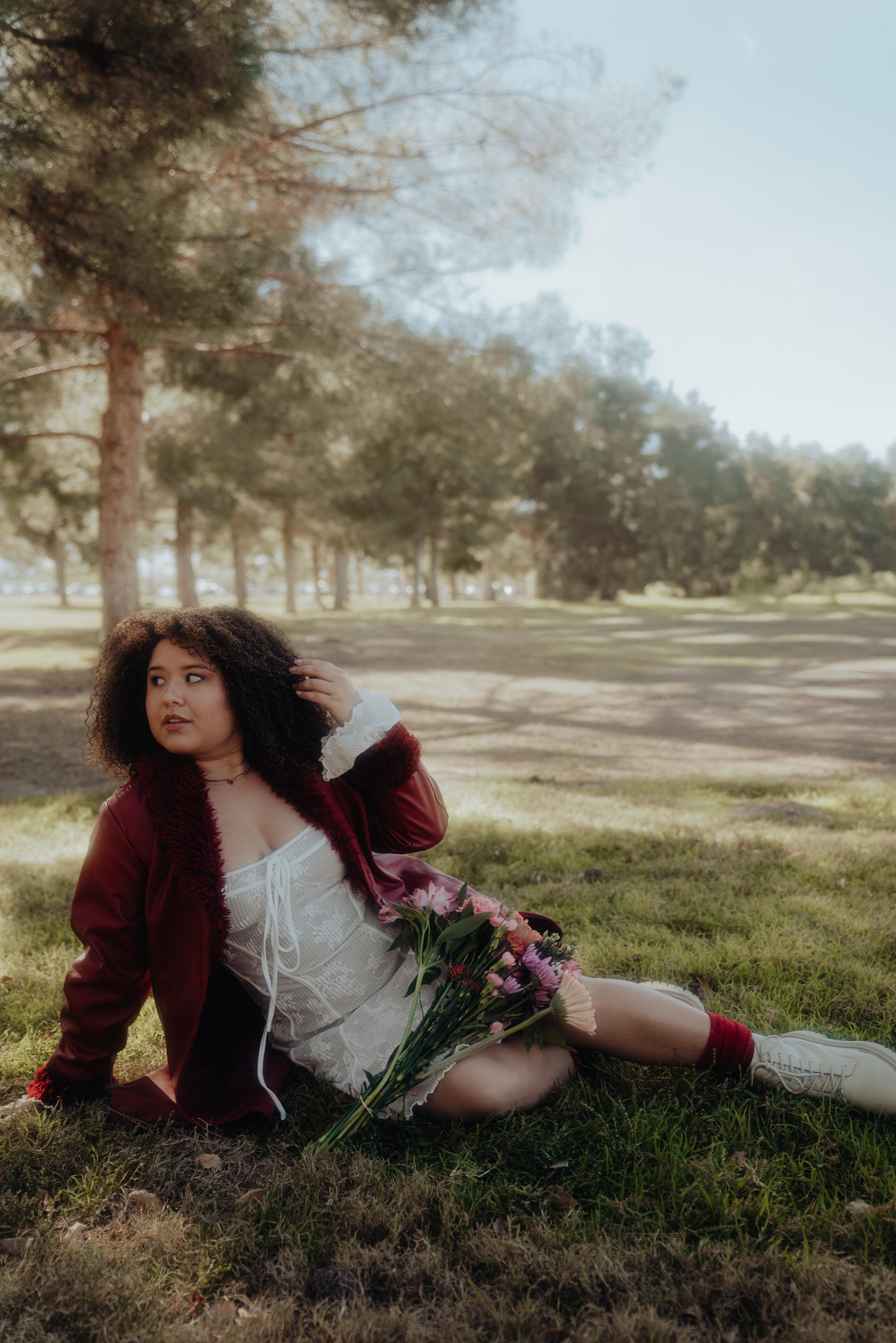 A woman with curly hair sitting on the grass in a park during daytime, holding a bouquet of pink flowers, with trees and a blue sky in the background.