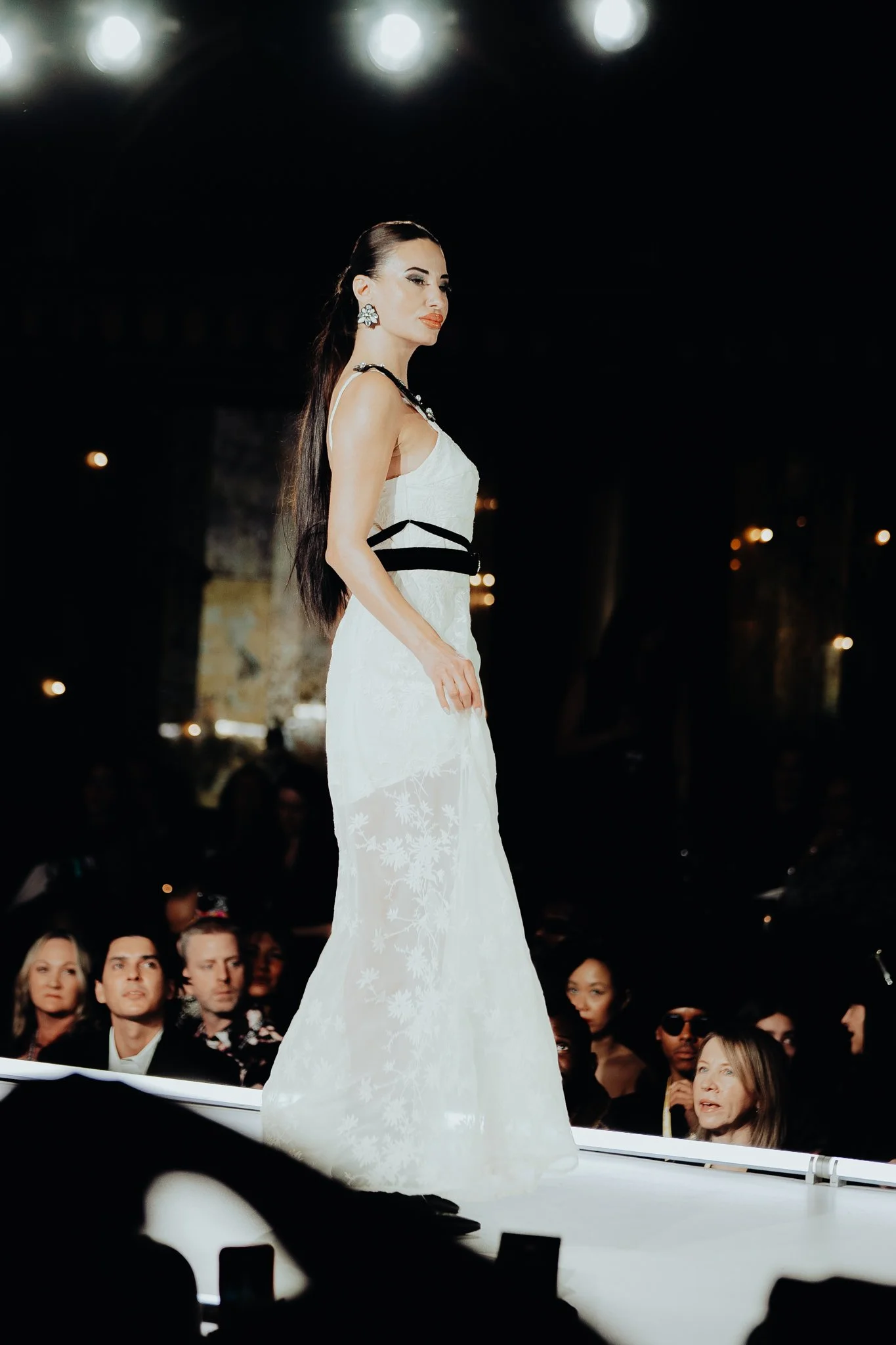 A woman in a white dress walking on a runway during a fashion show, with an audience seated on either side and bright lights overhead.