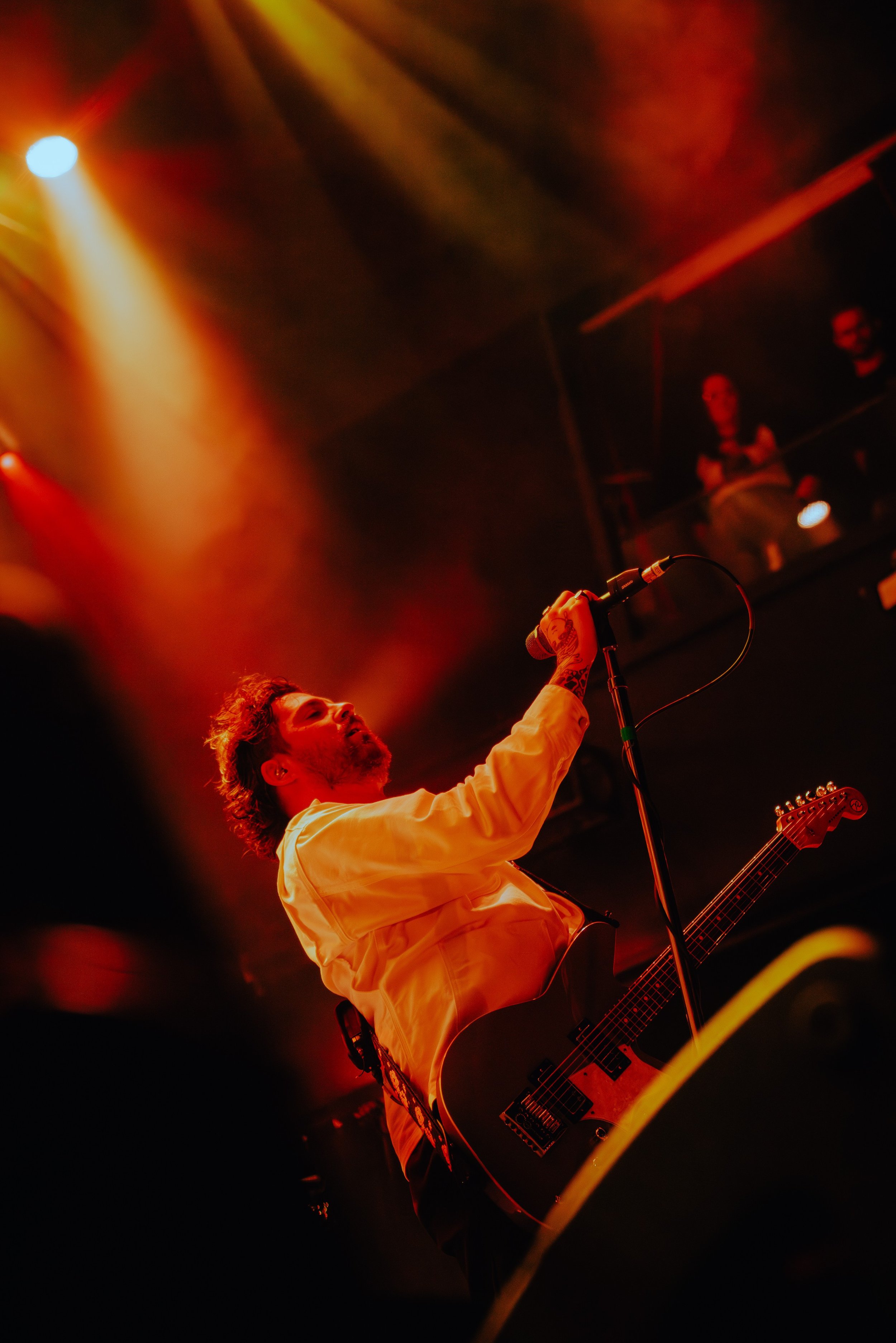 A male musician with curly hair and tattoos on his arm performing on stage with a guitar, illuminated by red and yellow stage lights, with a person in the background.