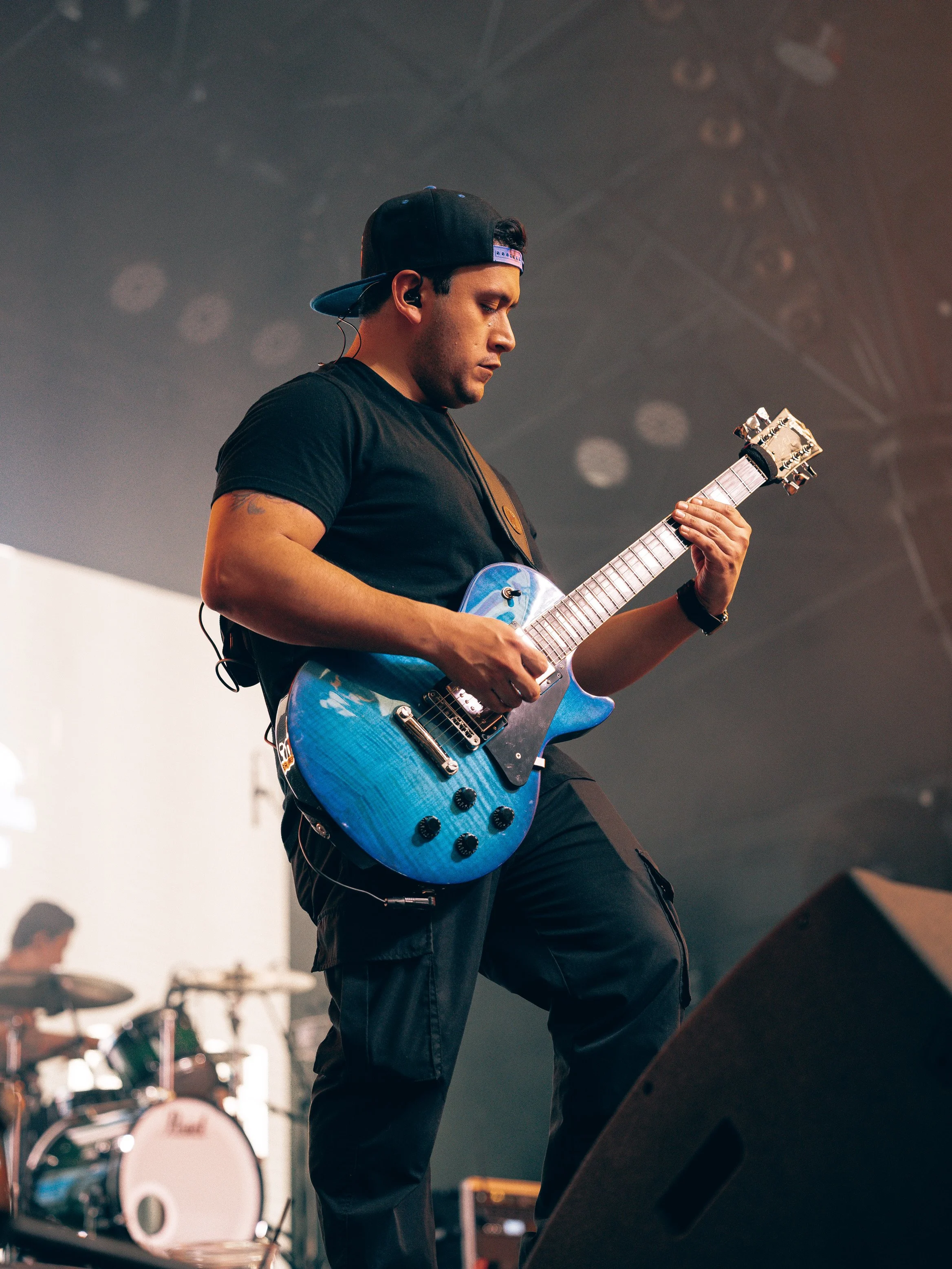 A man wearing a black cap backwards, black t-shirt, and wireless earbud, playing an electric guitar on stage with drums in the background.
