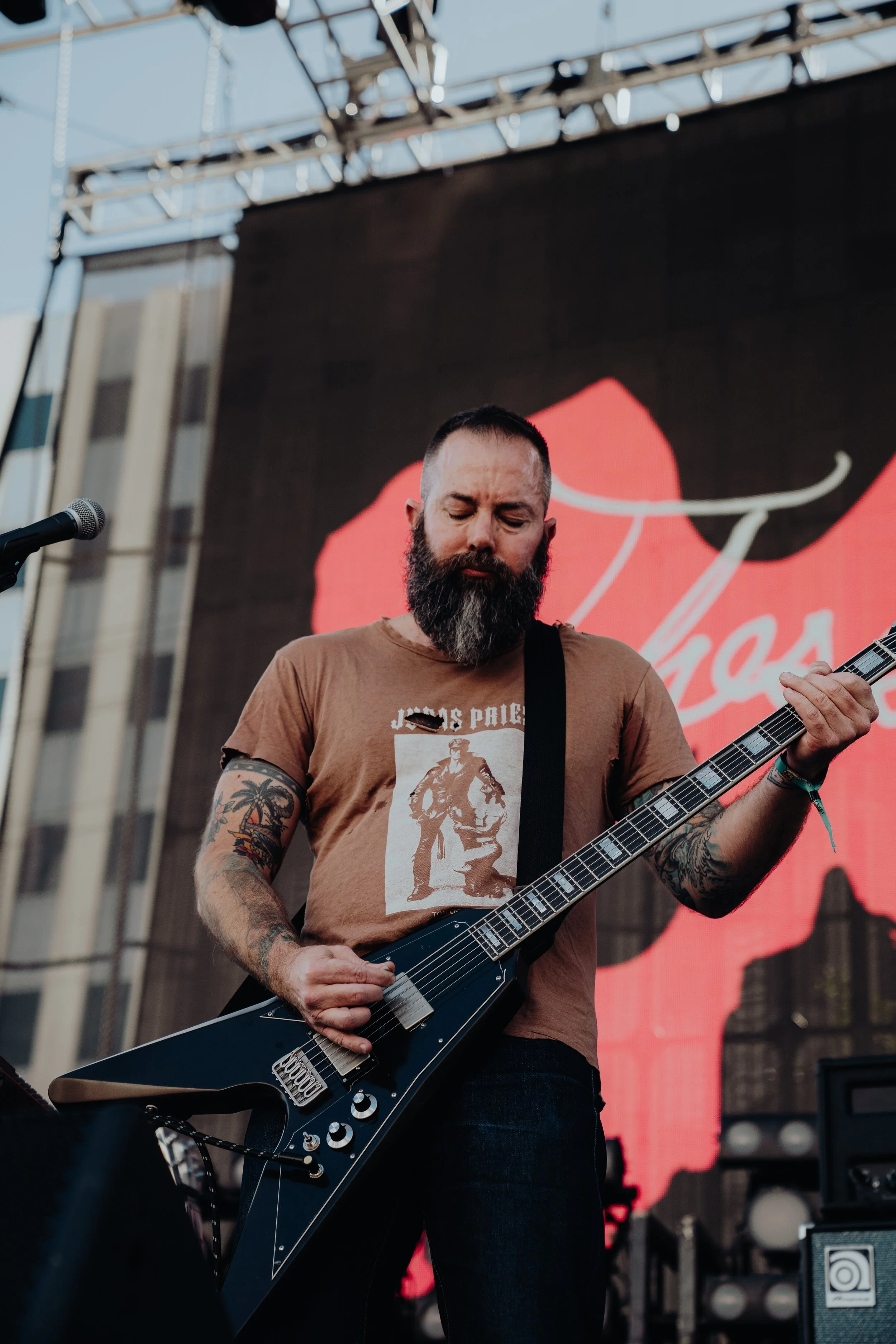 A bearded man playing an electric guitar on an outdoor stage with a large red and black backdrop.