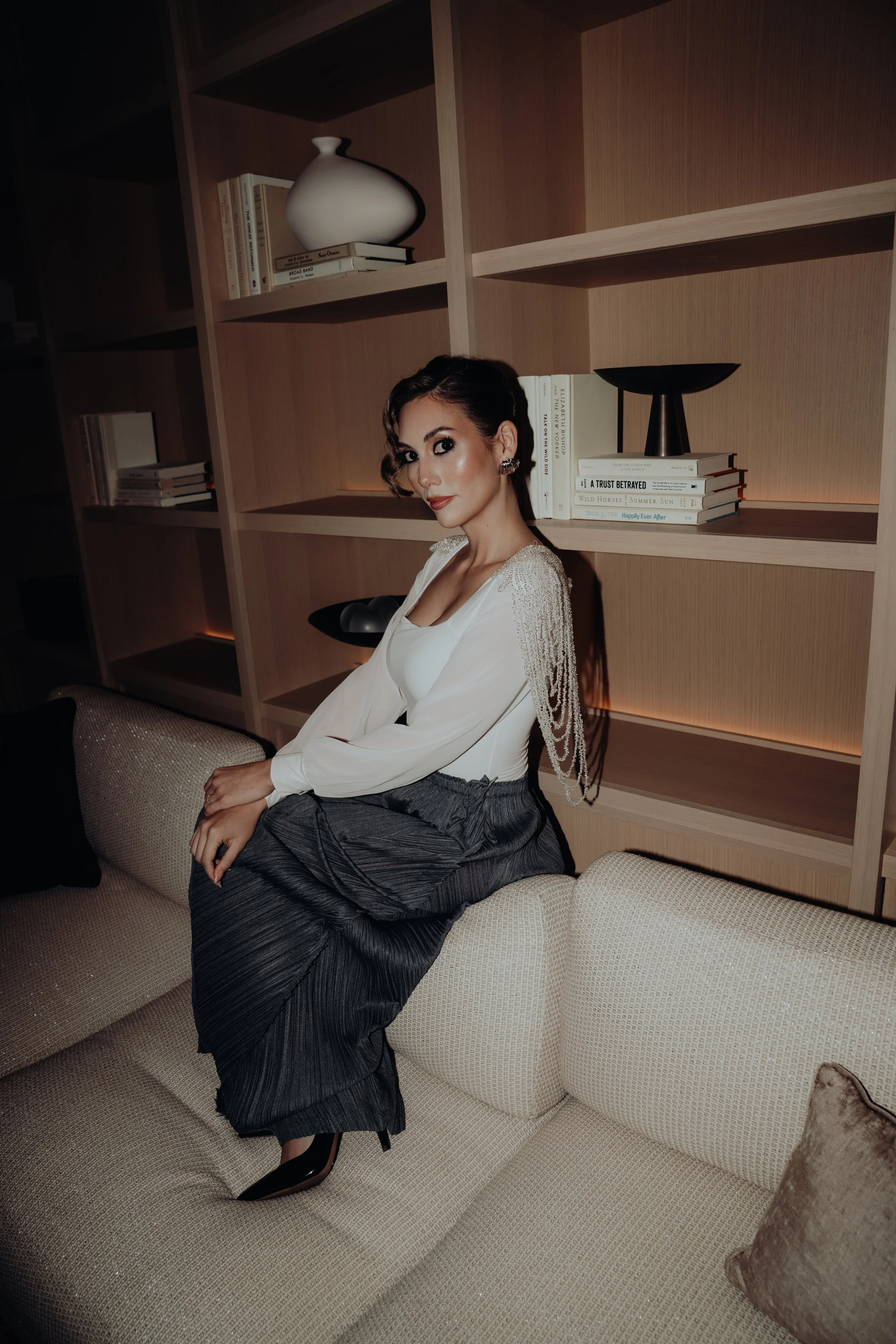 A woman with dark hair styled in an elegant updo, wearing a white top and dark pin-striped pants, sitting on a beige sofa in a room with a wooden bookshelf, modern decorative objects, and several books.