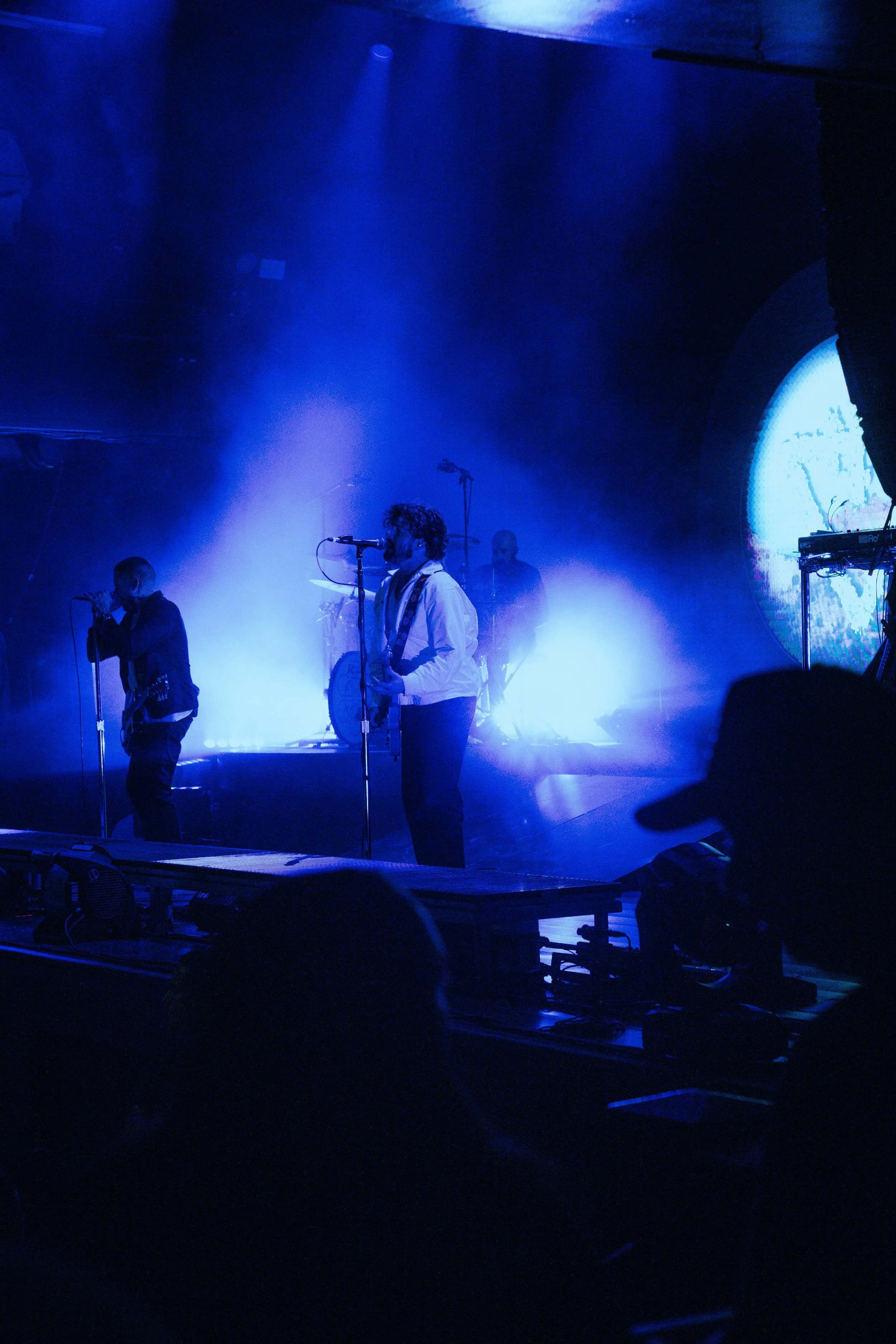 A musical band performing on a stage with blue lighting, including a singer, a saxophonist, and a drummer in the background, with a circular video projection on the right side of the stage.