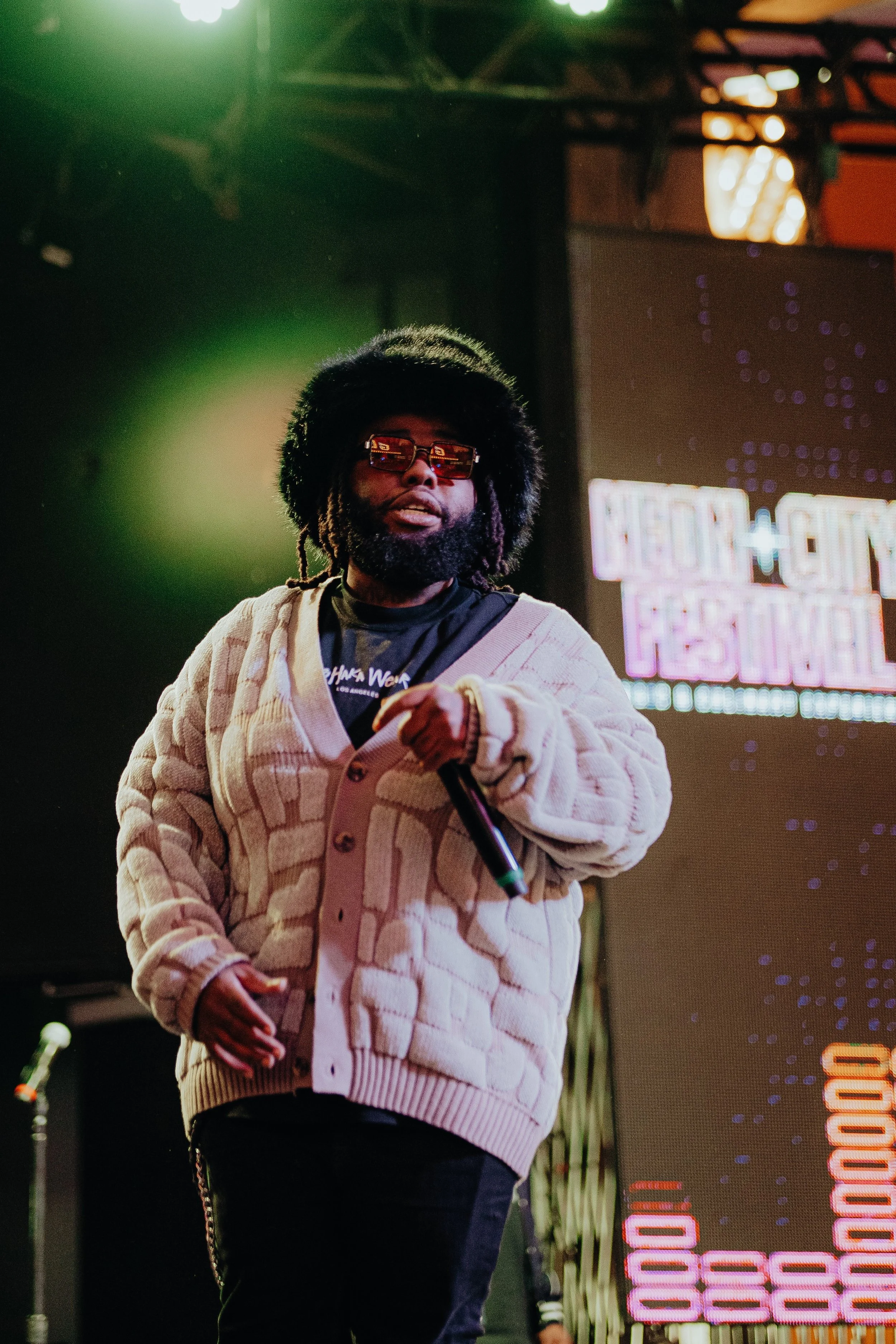 A man with a beard, wearing a beige jacket, red sunglasses, and a black shirt, holding a microphone on stage with bright, colorful lights and a digital display in the background.