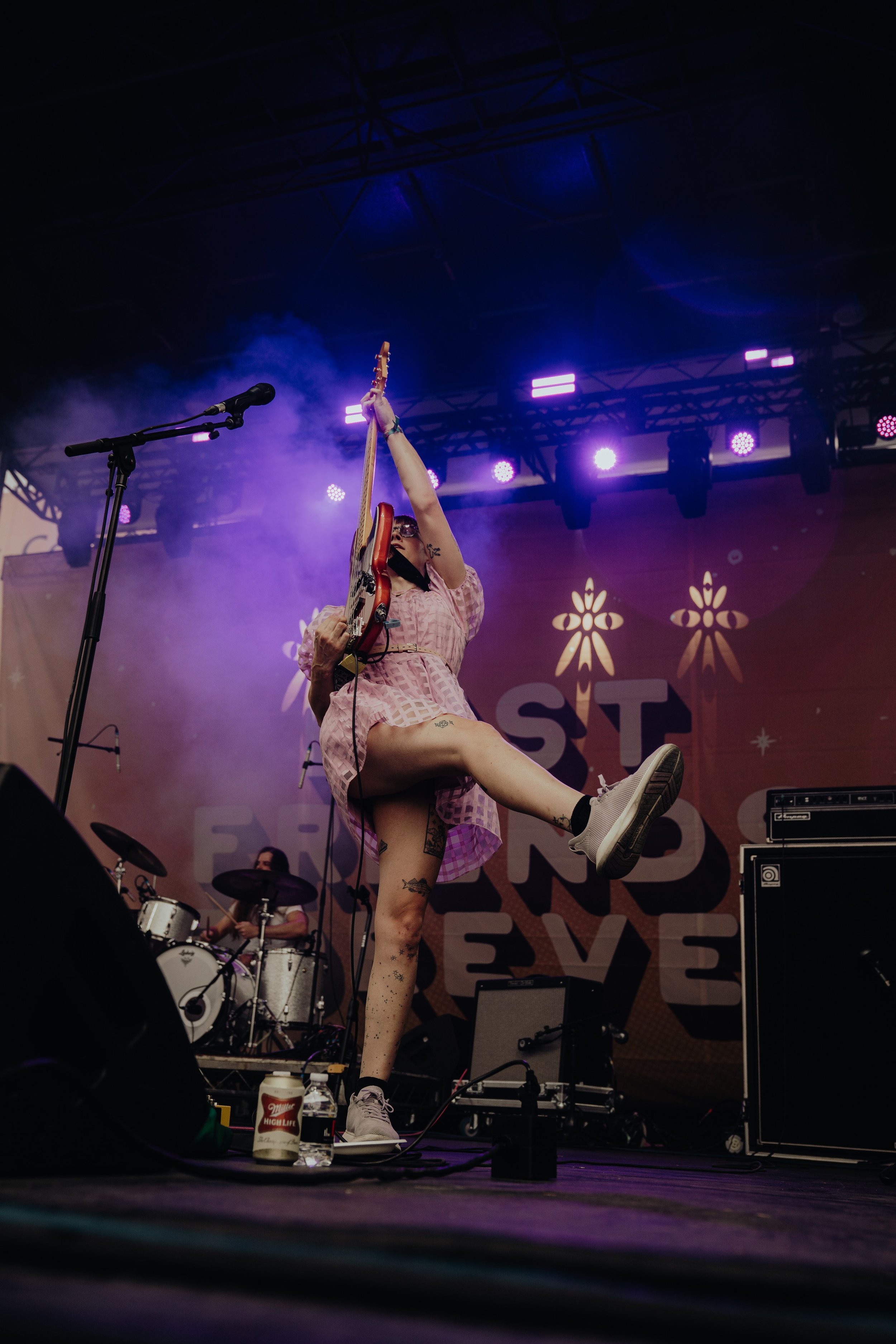 A performer on stage playing a guitar with one leg raised, wearing a pink checkered outfit and sneakers, with purple stage lights and a drum set in the background at a music festival.