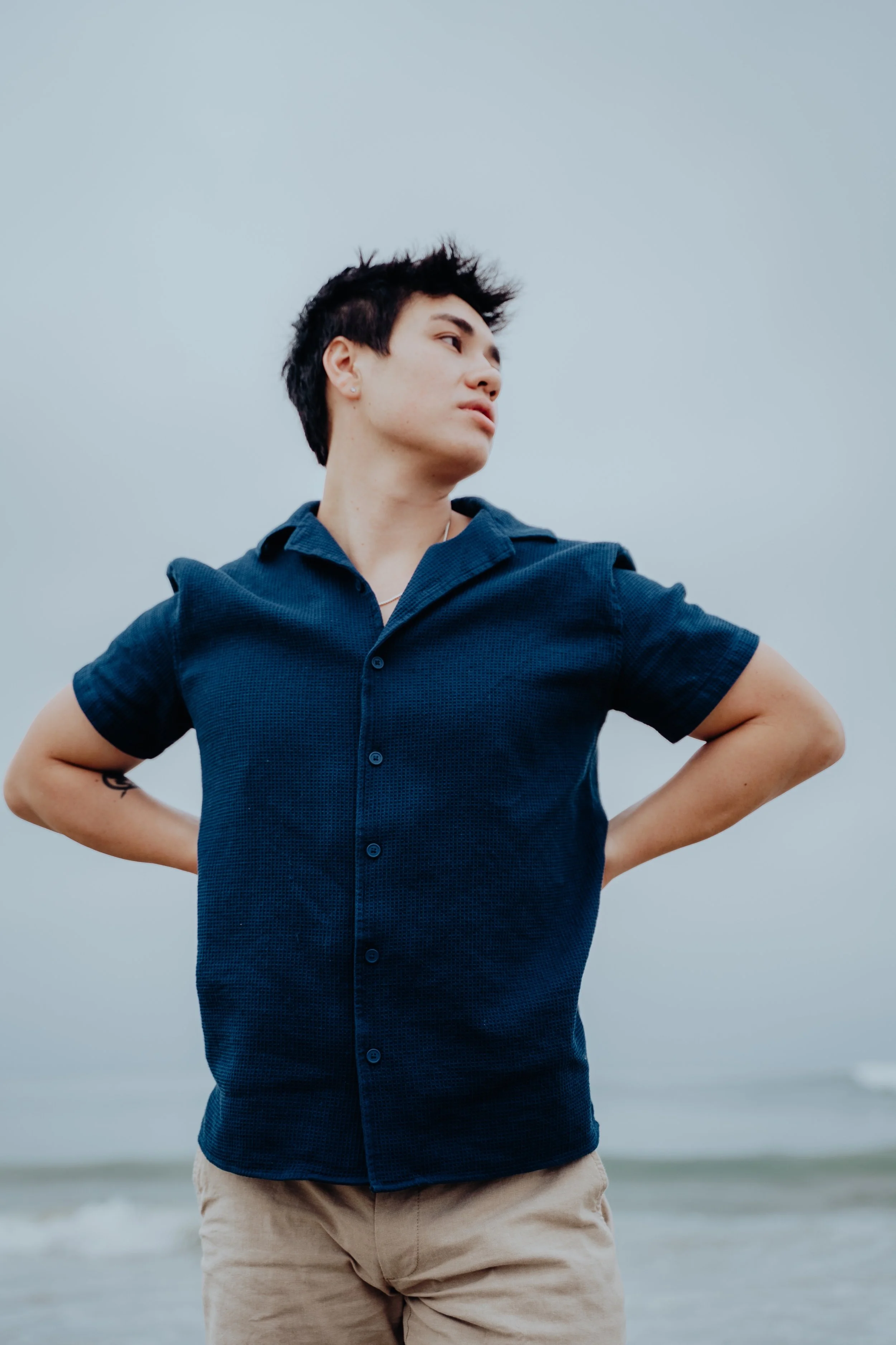 A young man with dark, tousled hair stands at the beach with his hands on his hips, looking to the side against a cloudy sky.