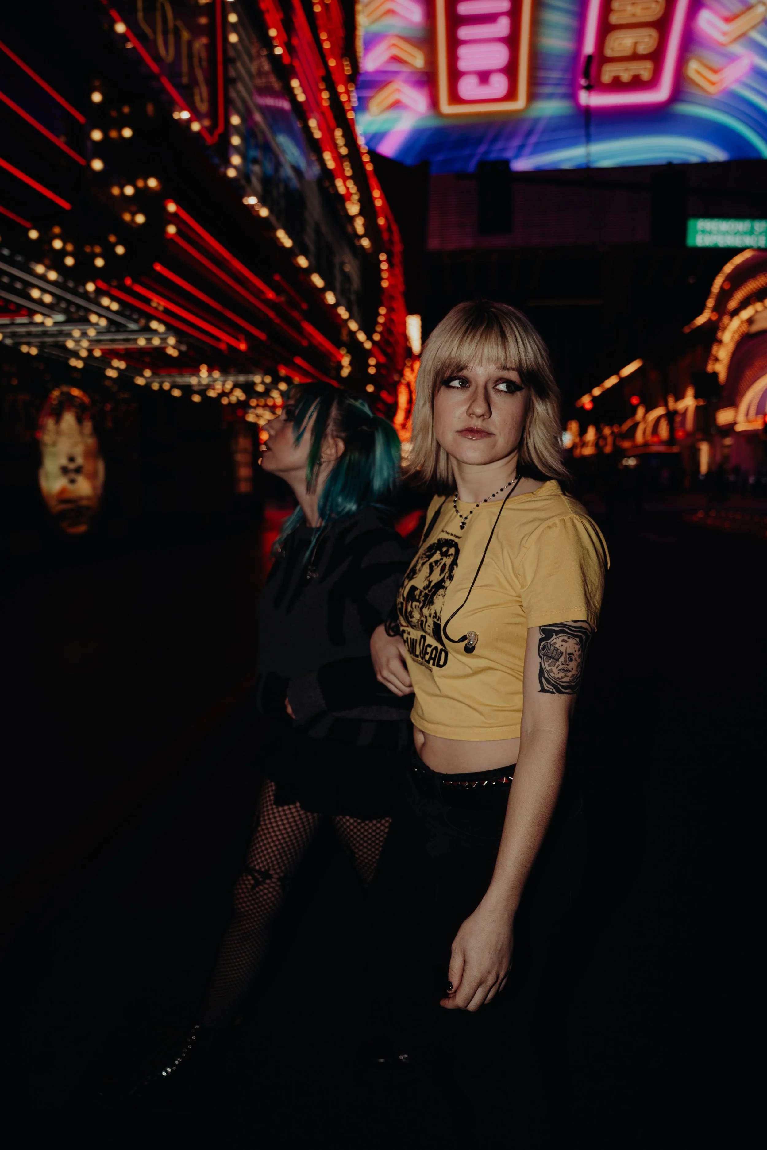 Two women standing in front of a brightly lit carnival or fair at night, with neon signs and colorful lights overhead. One woman wears a yellow crop top with tattoos on her arm, and the other woman has blue and green hair with a dark outfit.