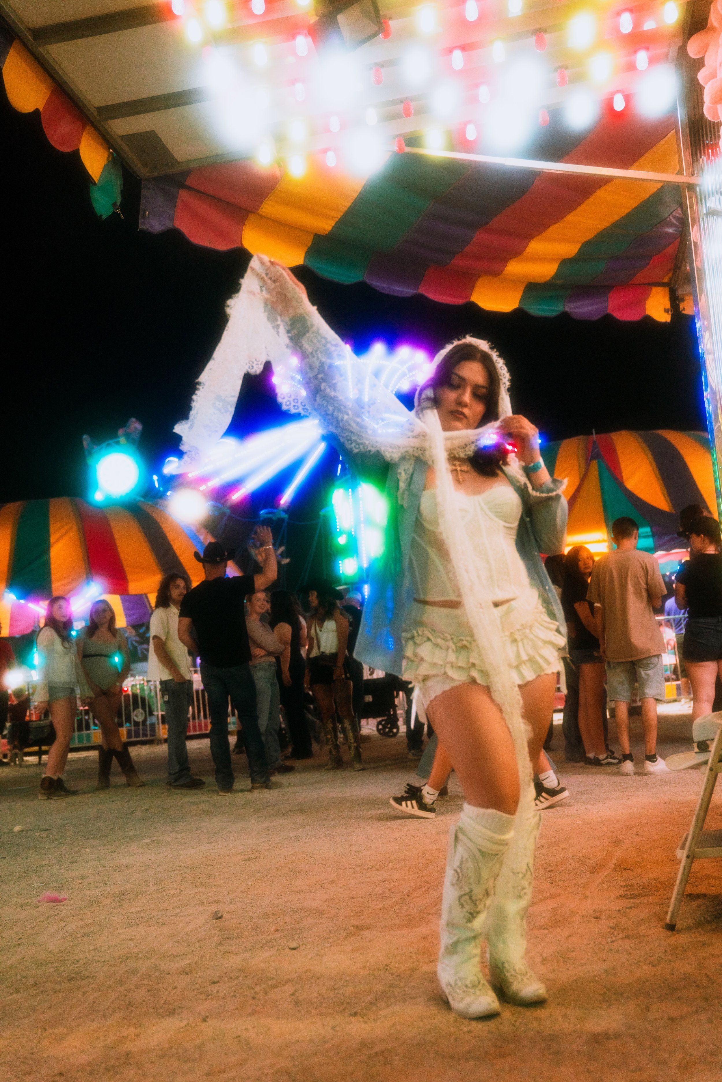 A woman dressed in a white lace dress with thigh-high stockings and sneakers, adjusting a lace veil at a carnival or fair with a colorful striped canopy, bright lights, and a crowd of people in the background.