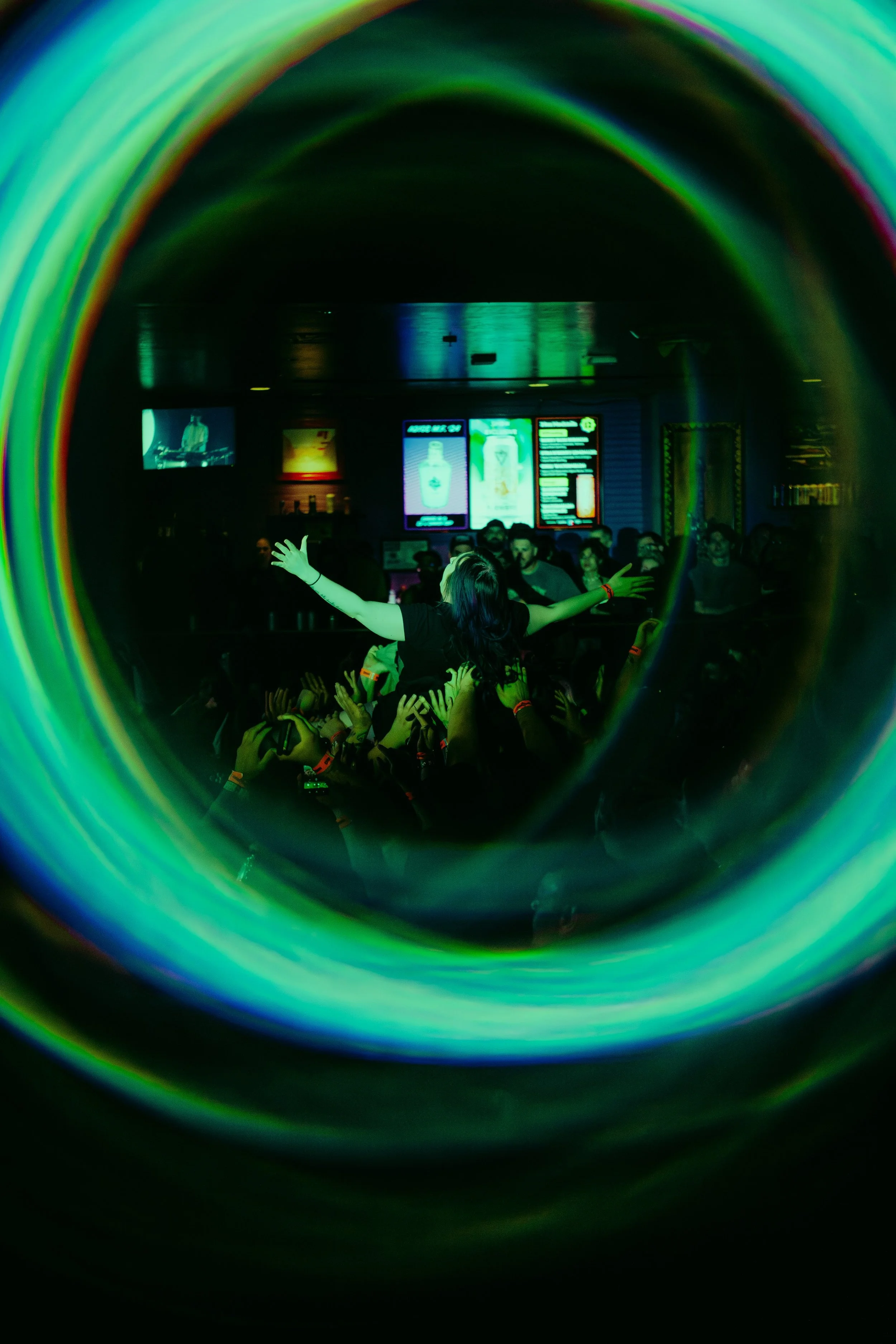 A woman performing on stage at a concert, with her arms outstretched, seen through a colorful, circular light reflection. The audience is visible, with some people taking photos or recording, and a large screen in the background displays a bottle and