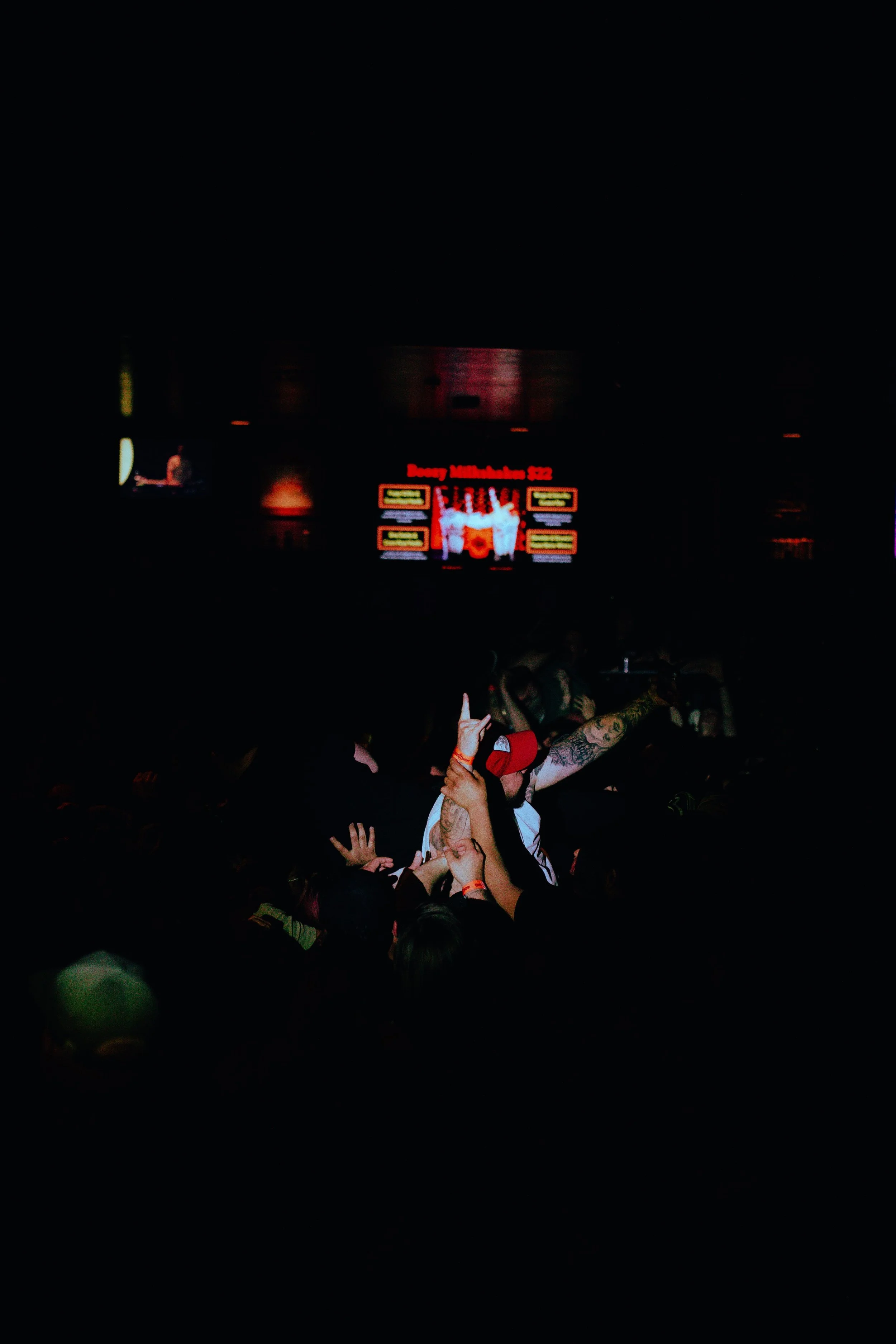 A dark concert venue with a group of people in the foreground, raising their hands and taking photos, and a screen in the background displaying a brightly lit image of a person. The screen reads 'Benny Muthalal 52.'