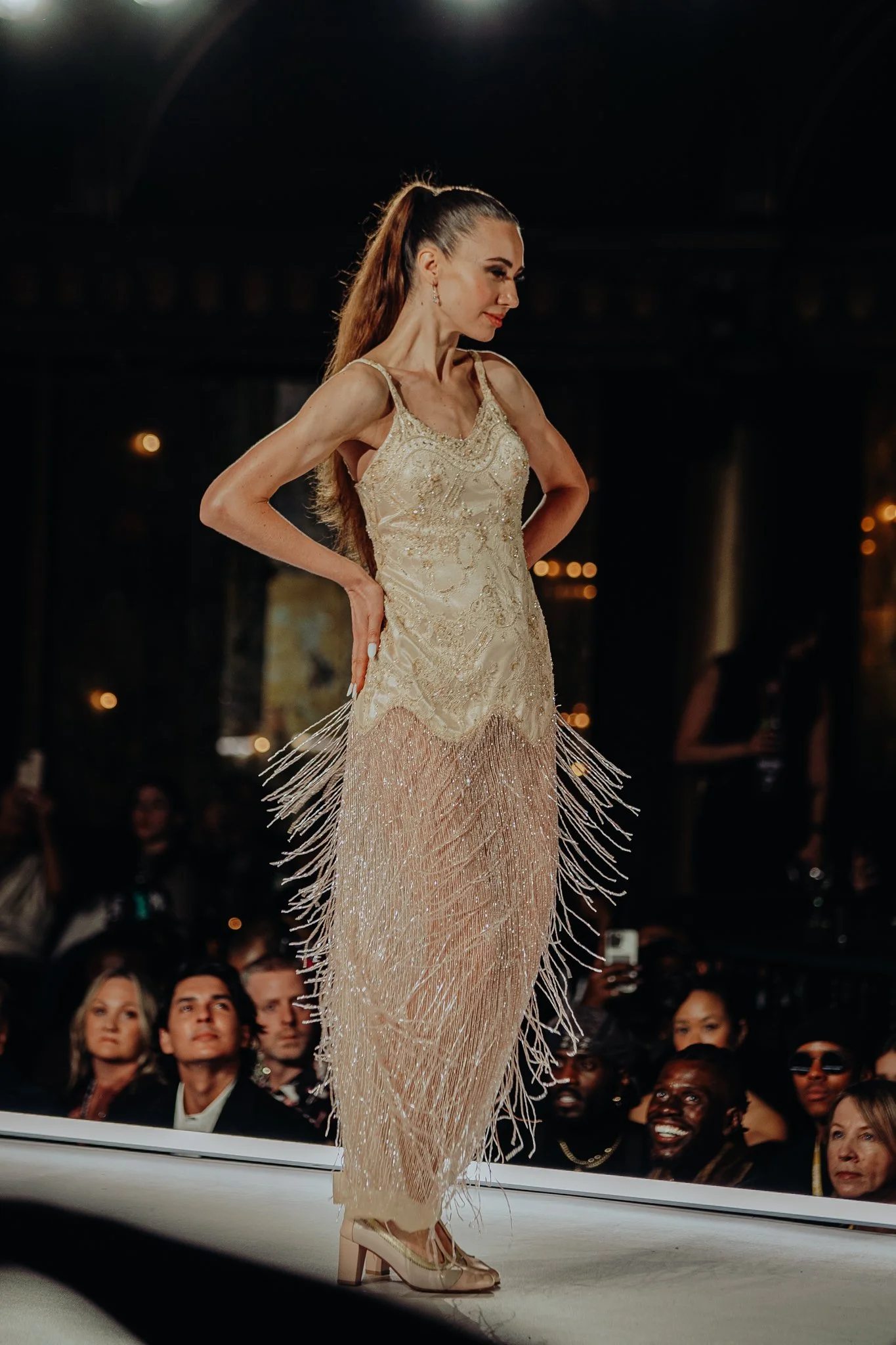 Model walking runway in a gold dress with fringe details and gold heels at a fashion show, with an audience watching.