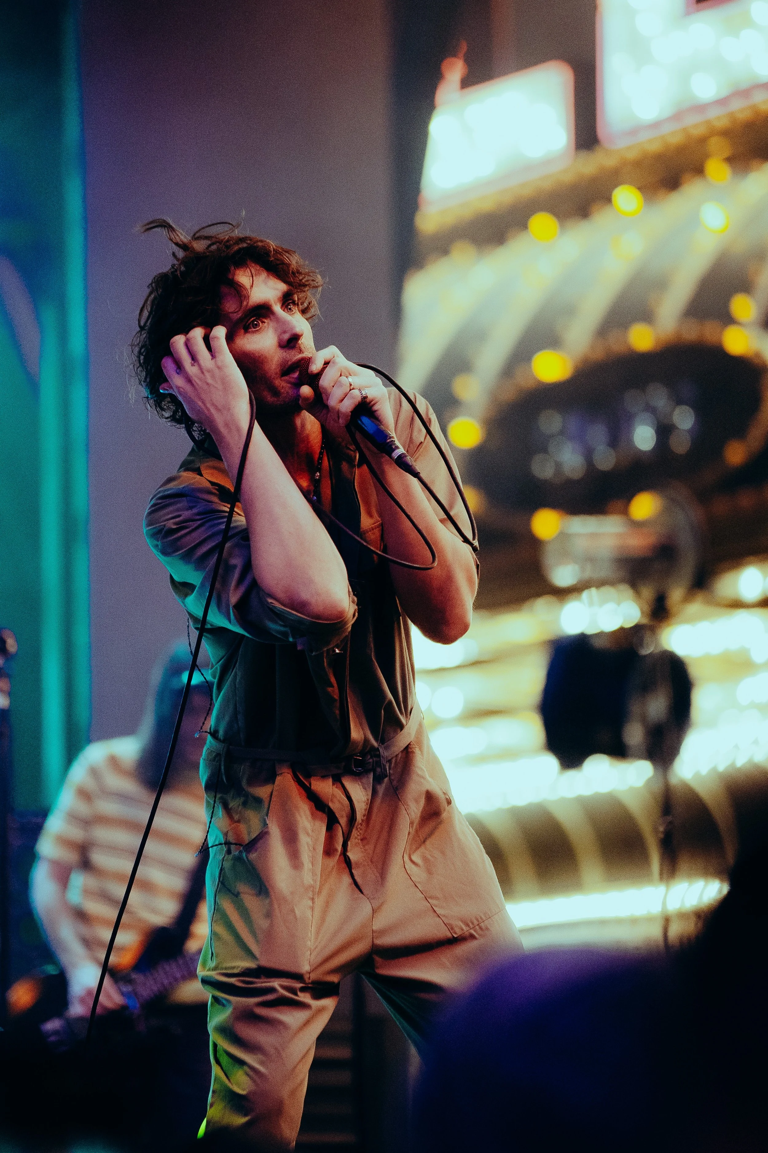 A man with curly hair holding a microphone in one hand and covering his ear with the other, standing in a neon-lit entertainment venue with a blurry background.