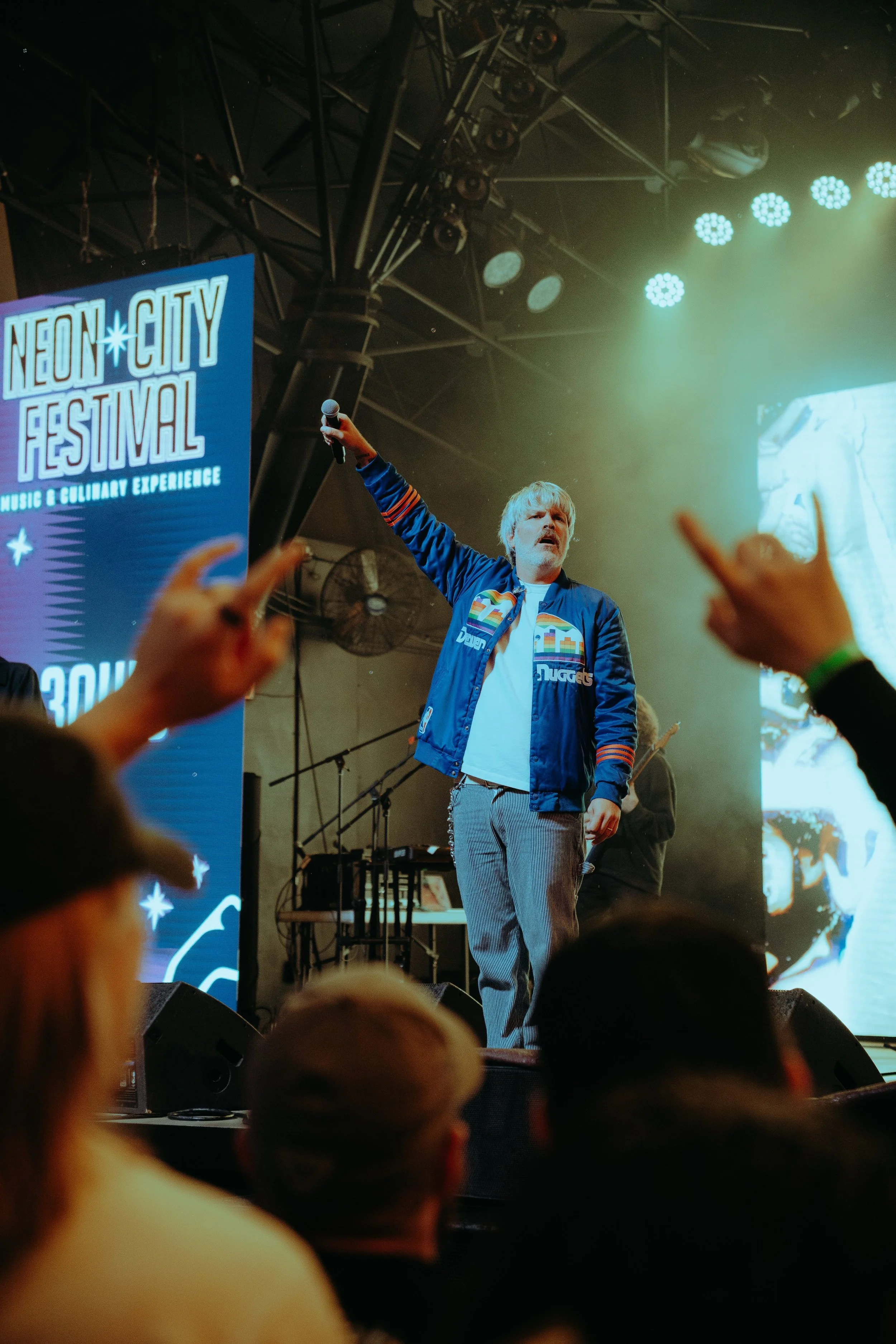Person on stage holding a microphone at the Neon City Festival, with a large screen and stage lights in the background, and an audience watching.
