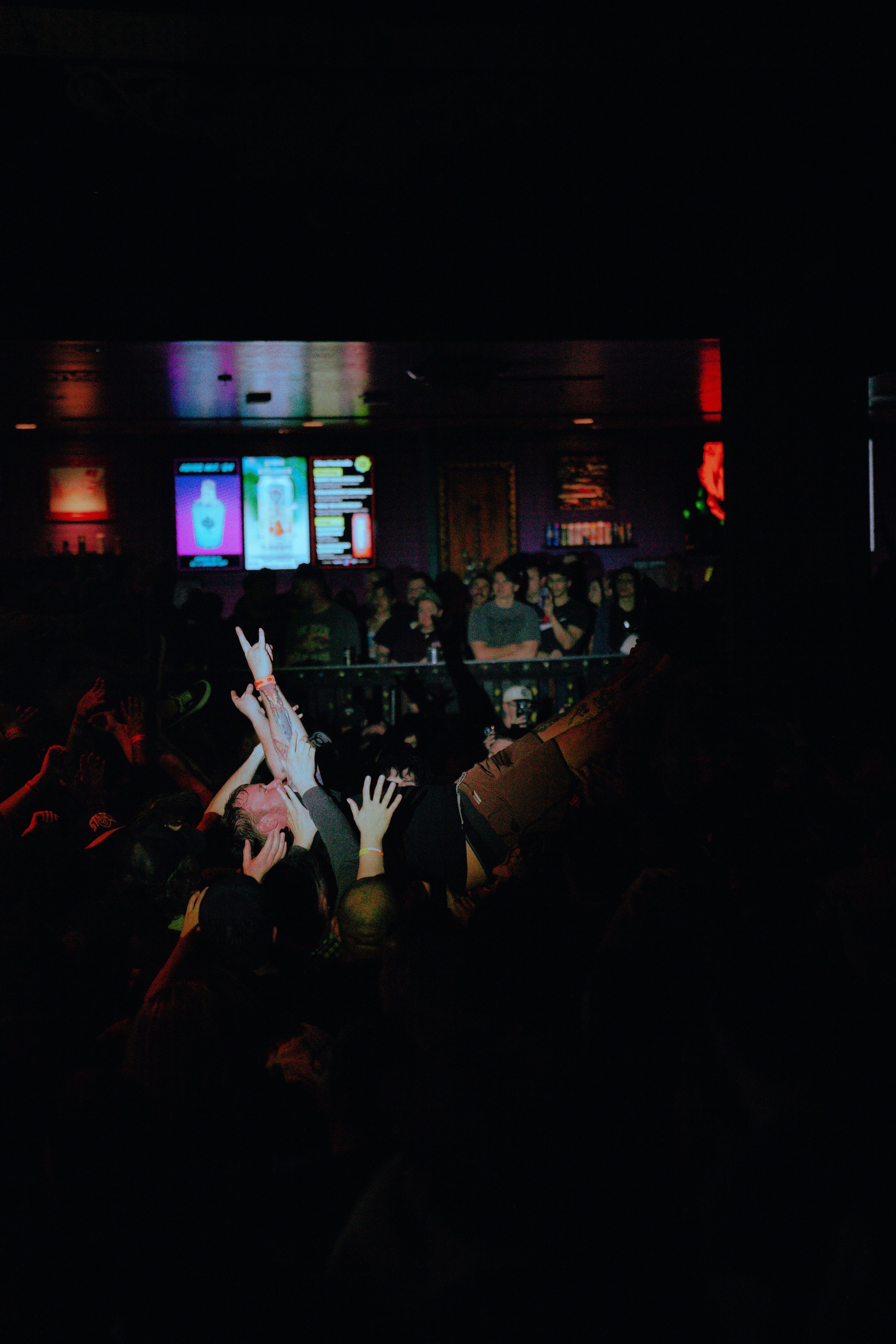 A crowded concert or nightclub scene with people raising their hands and dancing in front of a stage with illuminated screens and purple lighting.