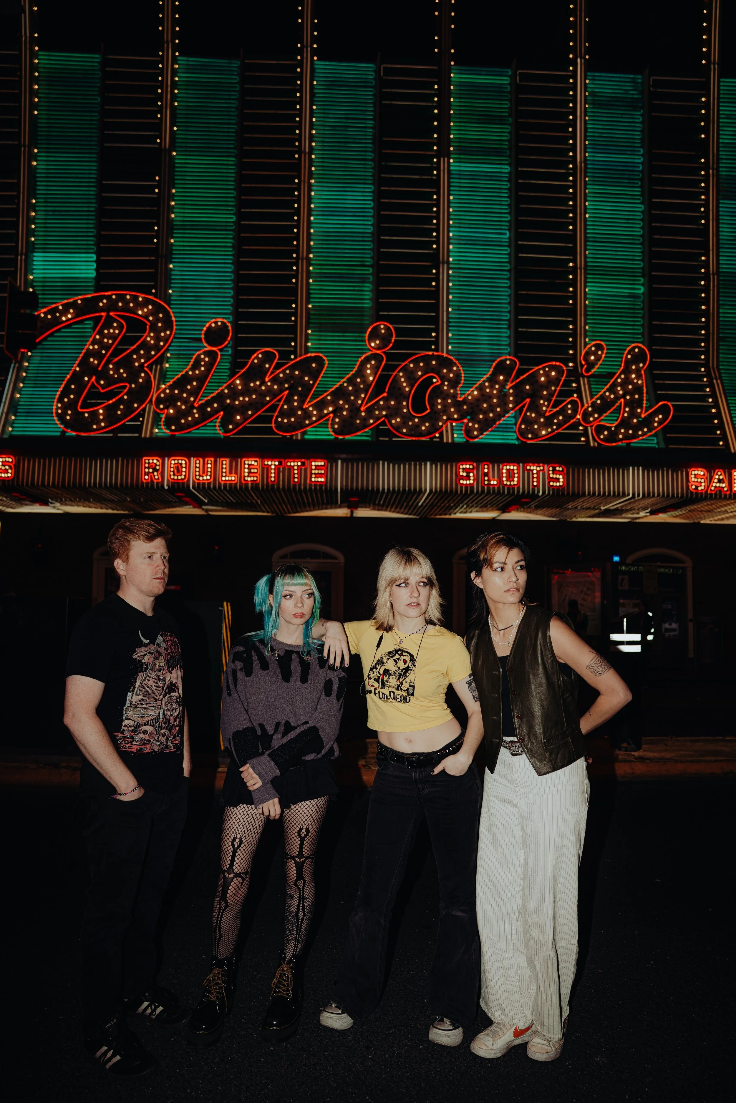Four young people standing in front of a brightly lit sign that says 'Binion's' with smaller signs reading 'Roulette,' 'Slots,' and 'S'. The group consists of two women and two men, dressed in casual and alternative styles, with colorful hair and tat