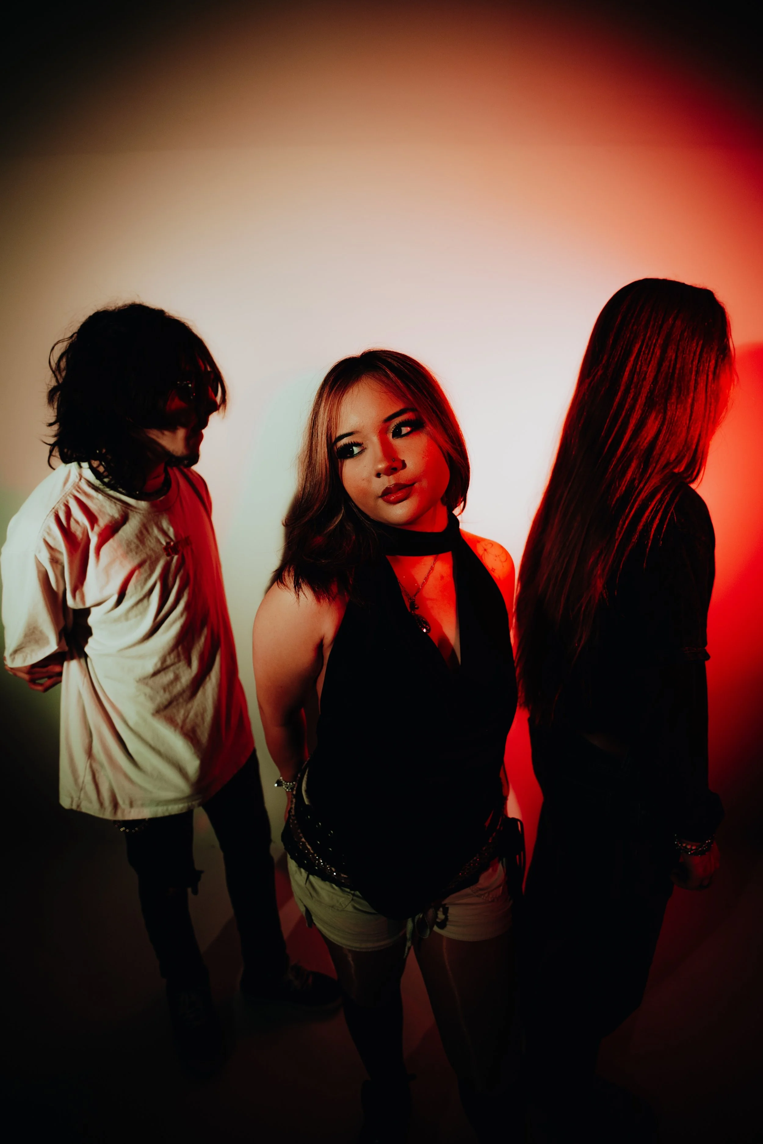 Group of three women standing in a dimly lit room with red lighting. The woman in the middle has shoulder-length brown hair, wearing a black top with a lace detail and shorts, looking to her right. The woman on the left has dark, wavy hair, wearing a