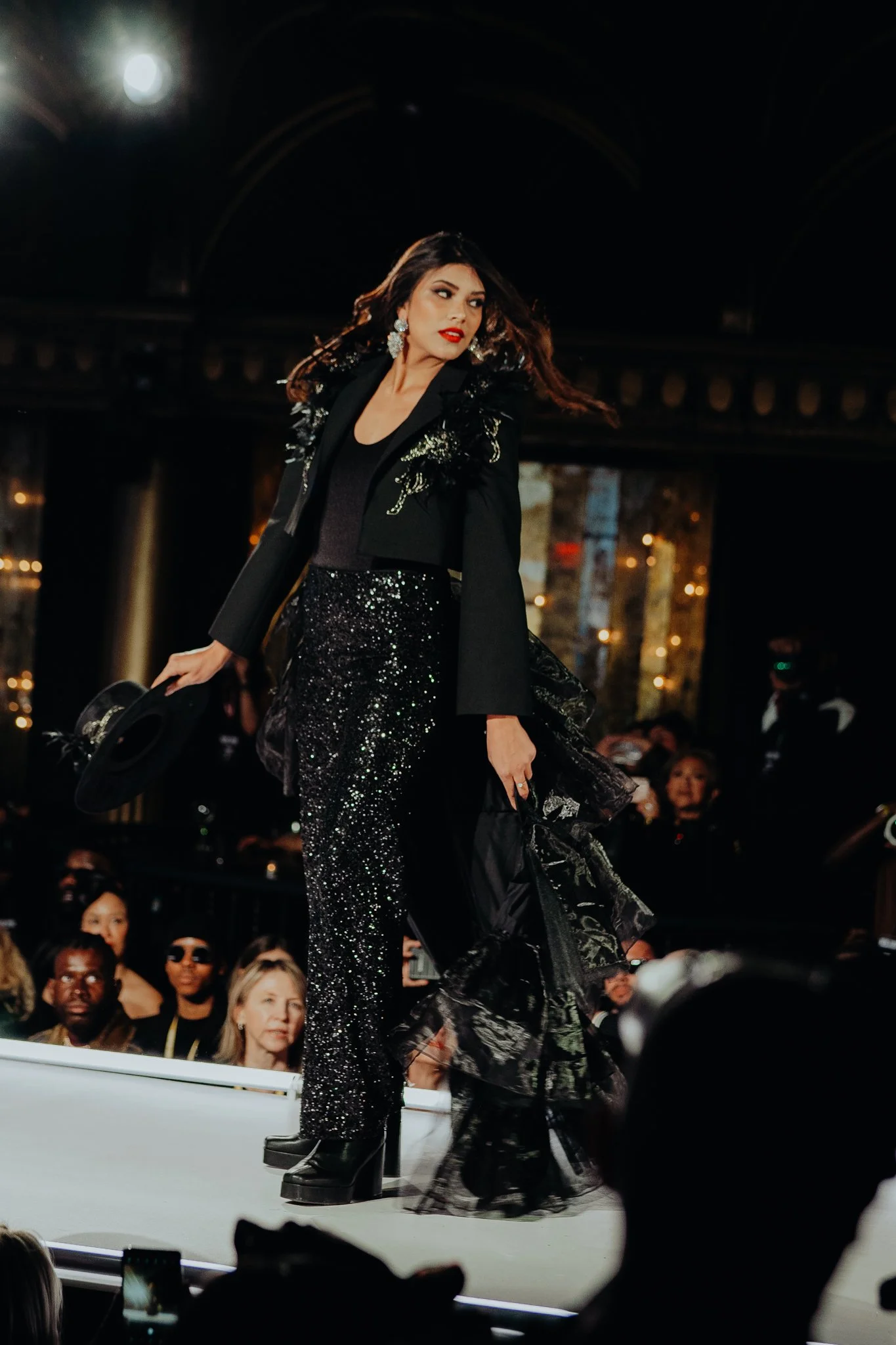 Fashion model walking on runway at night, dressed in black sequined outfit with large earrings, surrounded by an audience.