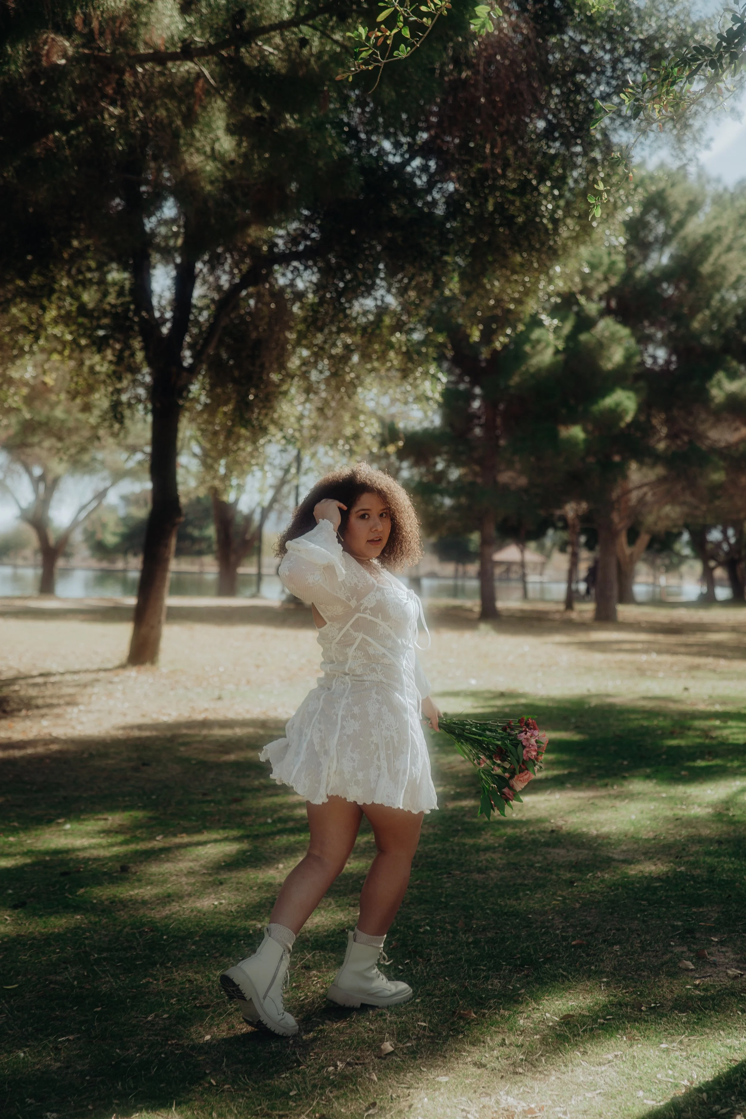 A woman outdoors wearing a white lacy dress, white boots, and holding a bouquet of pink flowers, standing in a park with trees and a body of water in the background.