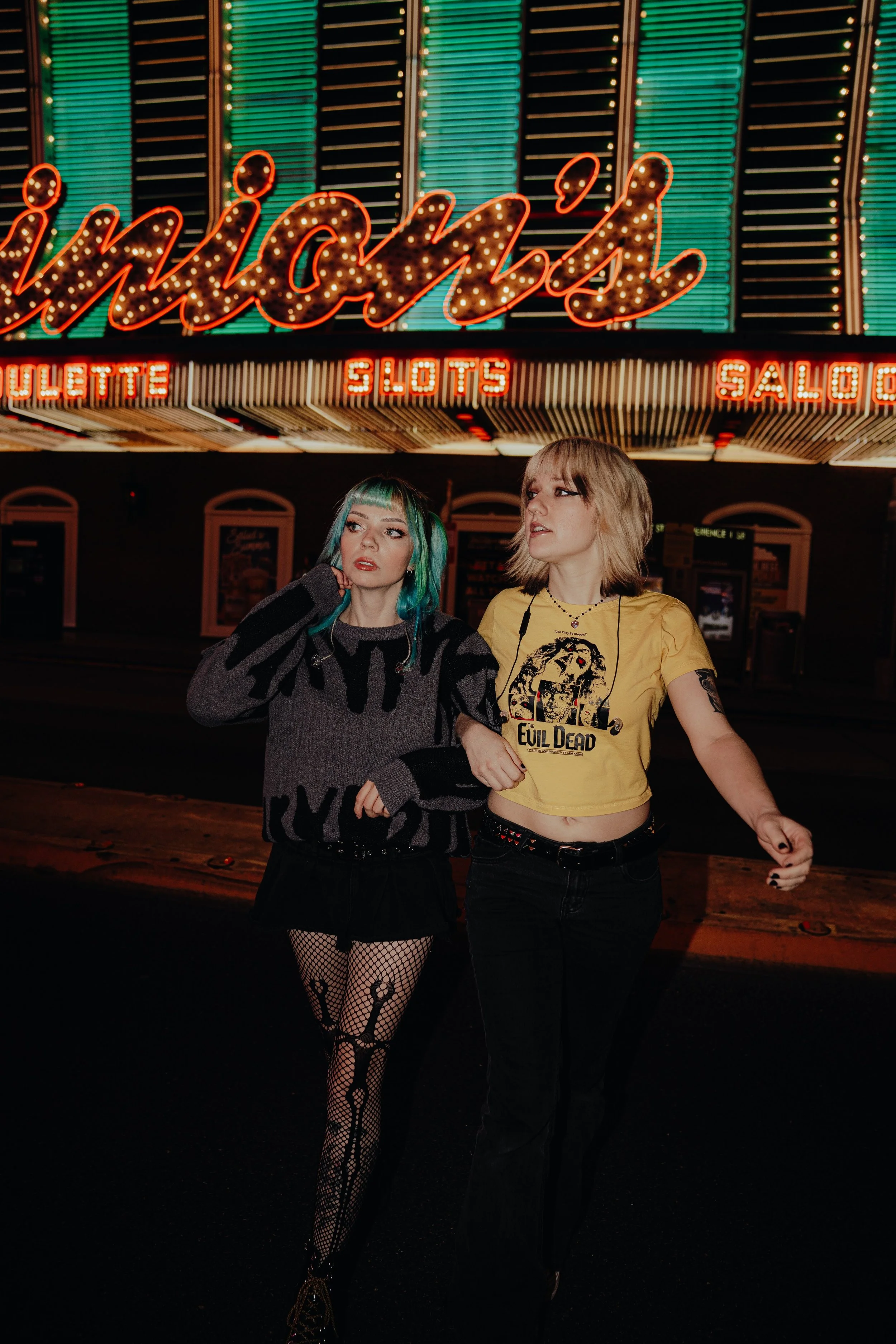 Two young women standing in front of a neon-lit casino marquee at night, one with teal hair and fishnet tights, the other with blonde hair and a The Evil Dead t-shirt.