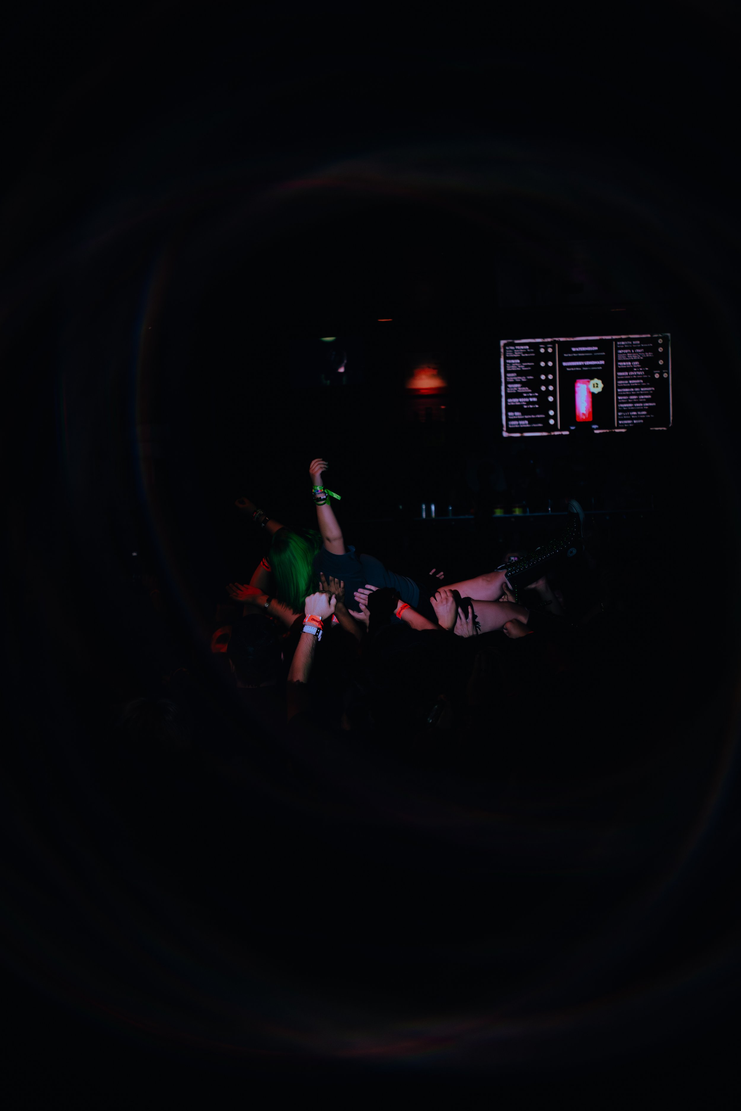 Dark scene with a digital screen displaying a menu or list, a person with green hair and wristbands dancing or enjoying music, arms raised in a concert or nightclub setting.