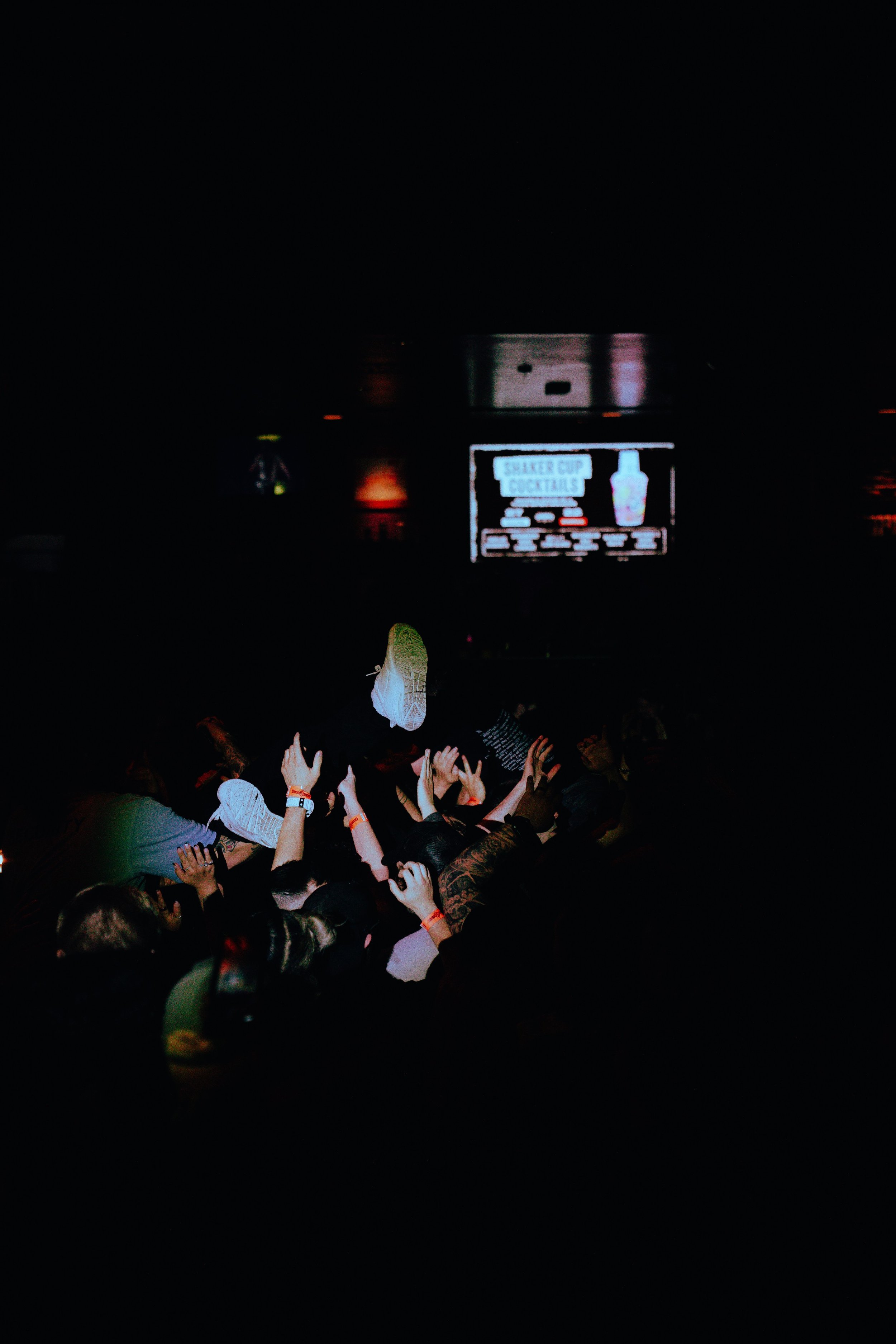Crowd of people reaching their hands towards a performer on stage at a concert, with a screen in the background displaying a cocktail advertisement for a shaker cup and cocktails.