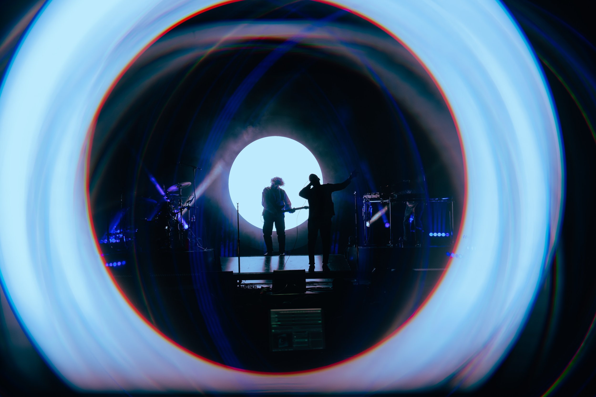 Silhouettes of musicians performing on stage, viewed through a colorful circular tunnel with lights and equipment visible.