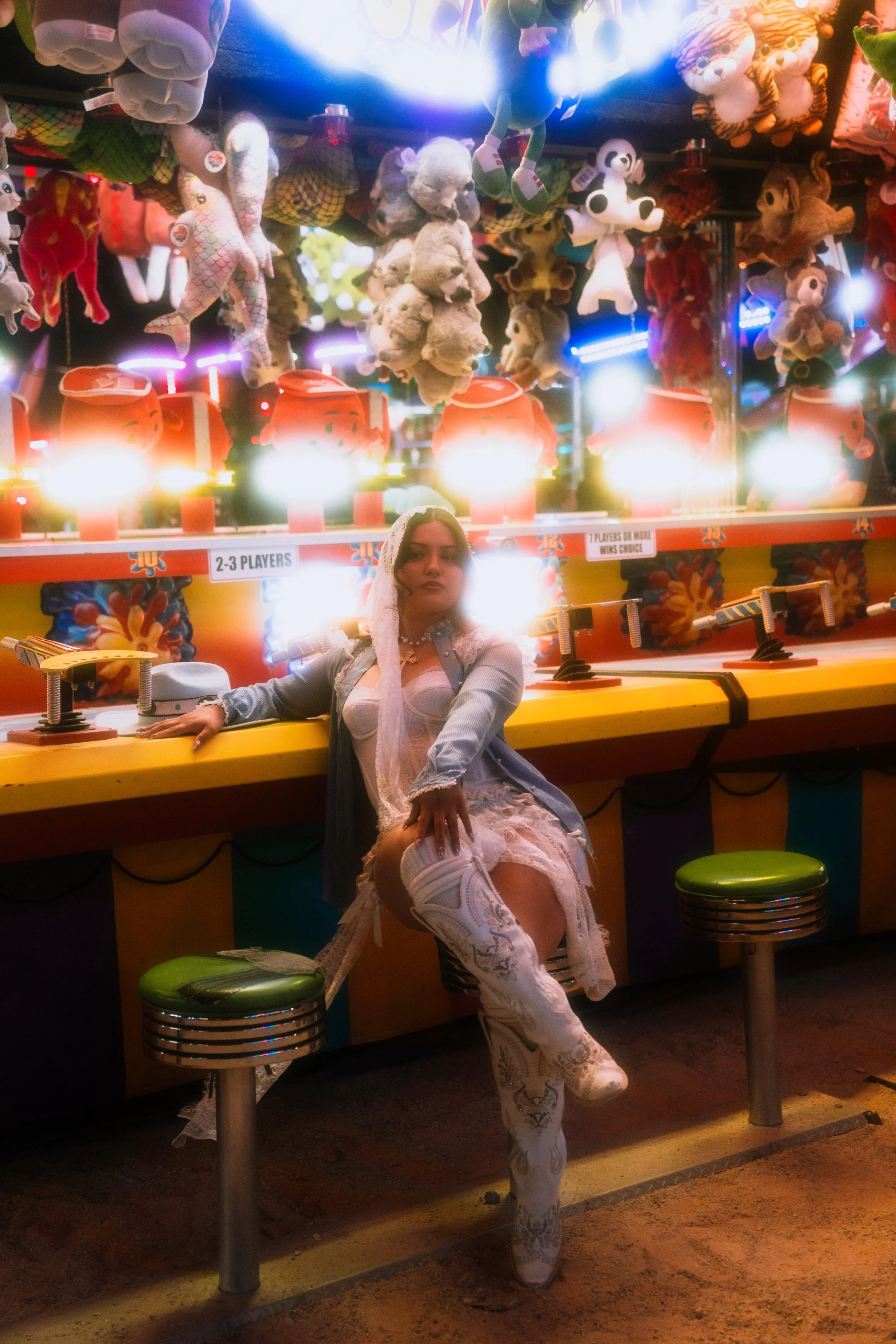 A woman dressed in a white lace dress and cowboy boots sitting in a carnival booth, with stuffed animal toys hanging behind her. She has long brown hair and is looking slightly to the side with a relaxed pose.