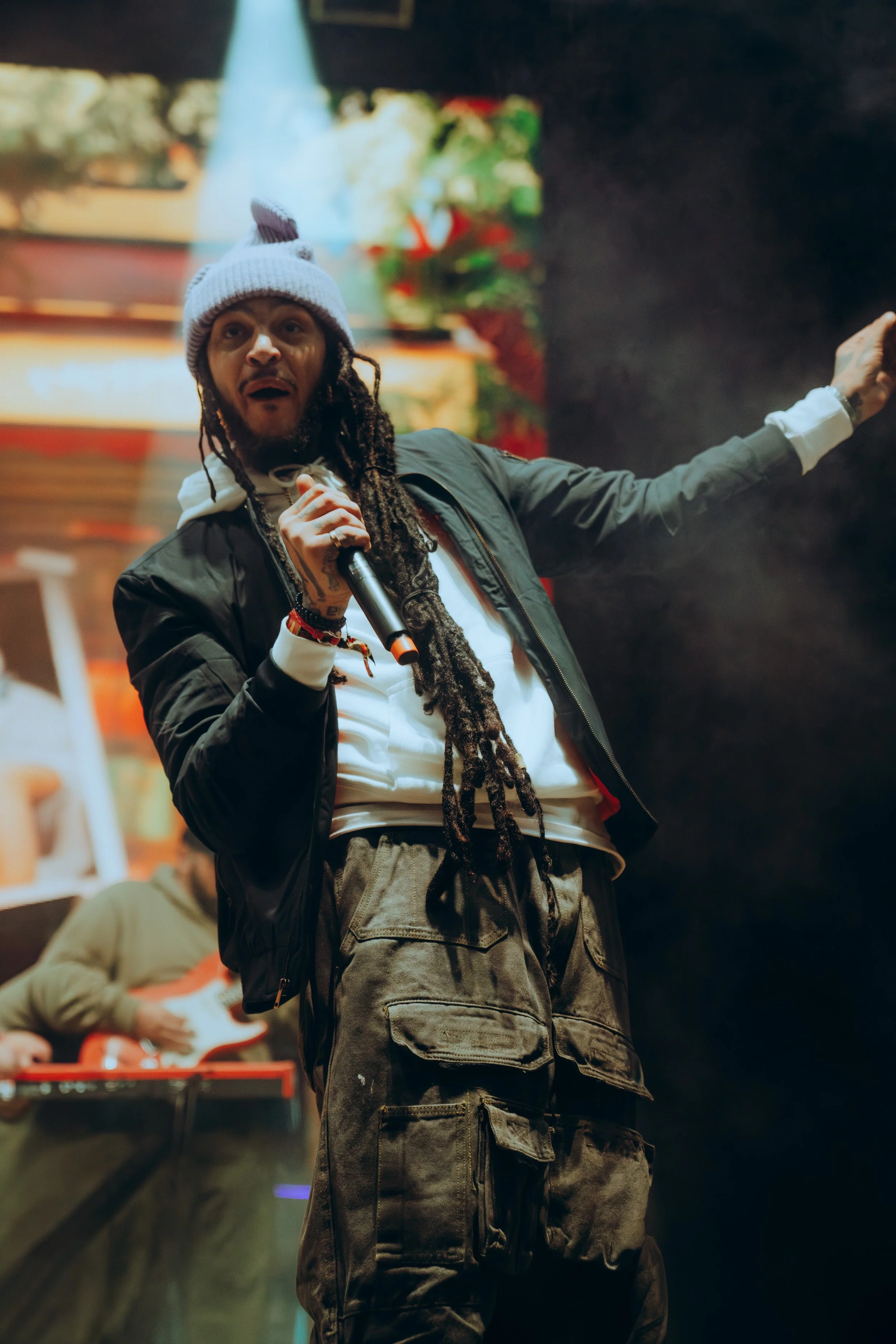 A male performer with dreadlocks wearing a gray beanie and black jacket singing into a microphone on stage, with another performer playing guitar in the background.