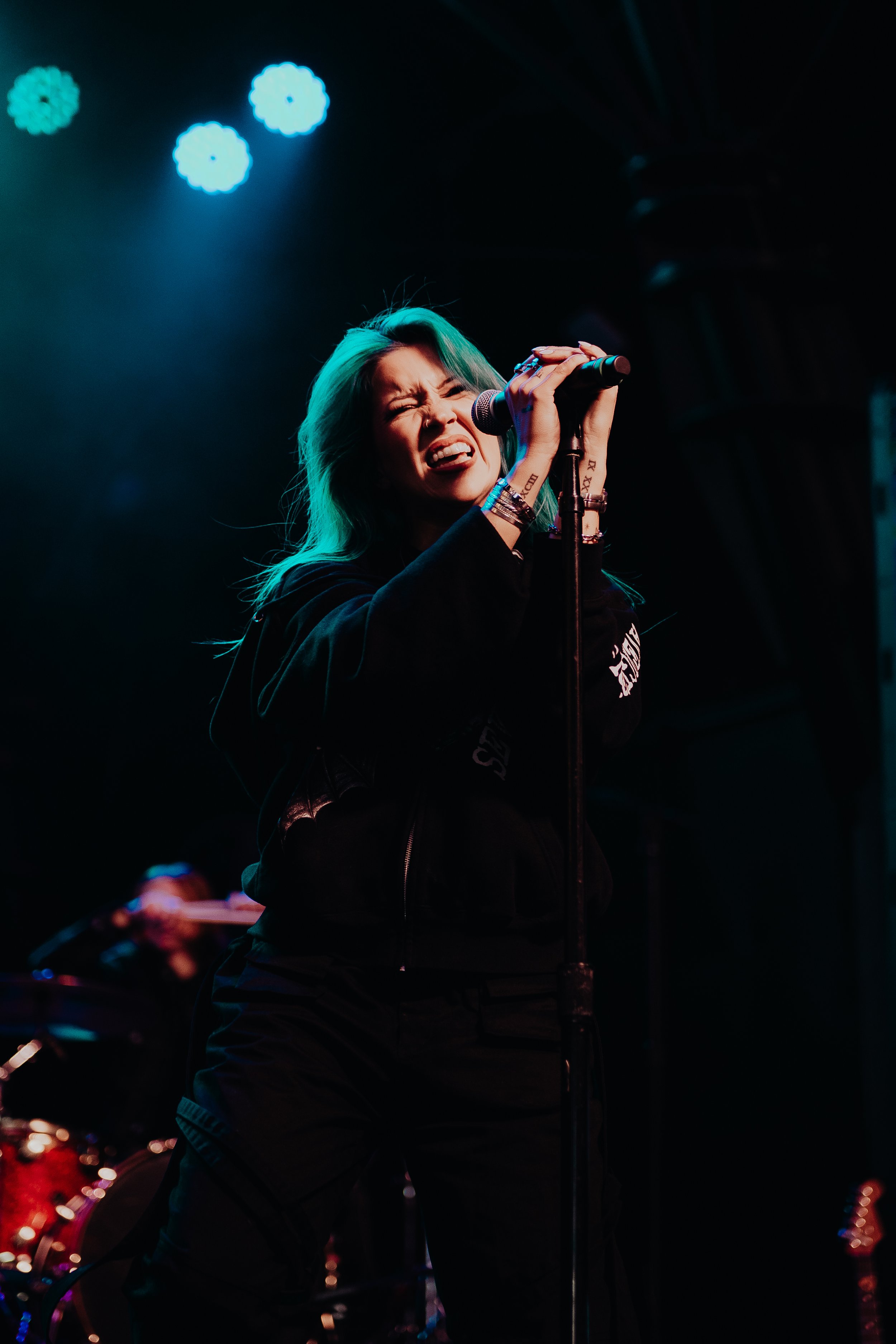 Female singer with turquoise hair singing into a microphone during a performance, with stage lights and drums in the background.