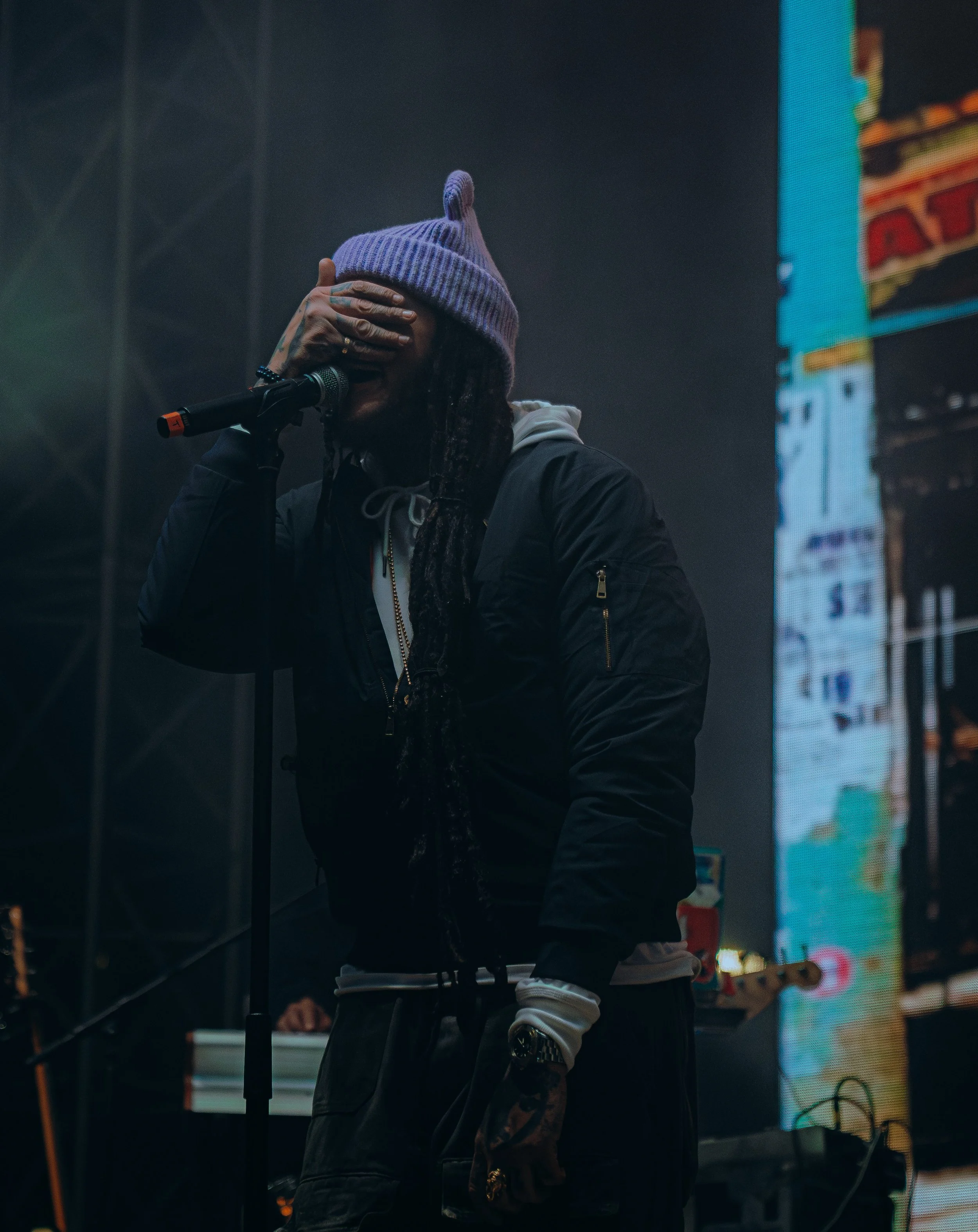 Musician performing on stage, wearing a purple knit hat, covering their face with one hand, with a microphone in front of them, illuminated by dark stage lighting.