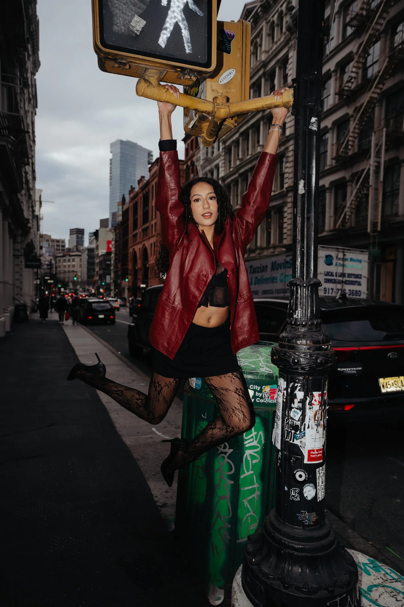 A woman in a red leather jacket, black fishnet stockings, and black underwear posing on a city street, holding onto a yellow pedestrian crossing signal with her arms raised above her head, one leg bent and foot resting on a green trash bin, with tall