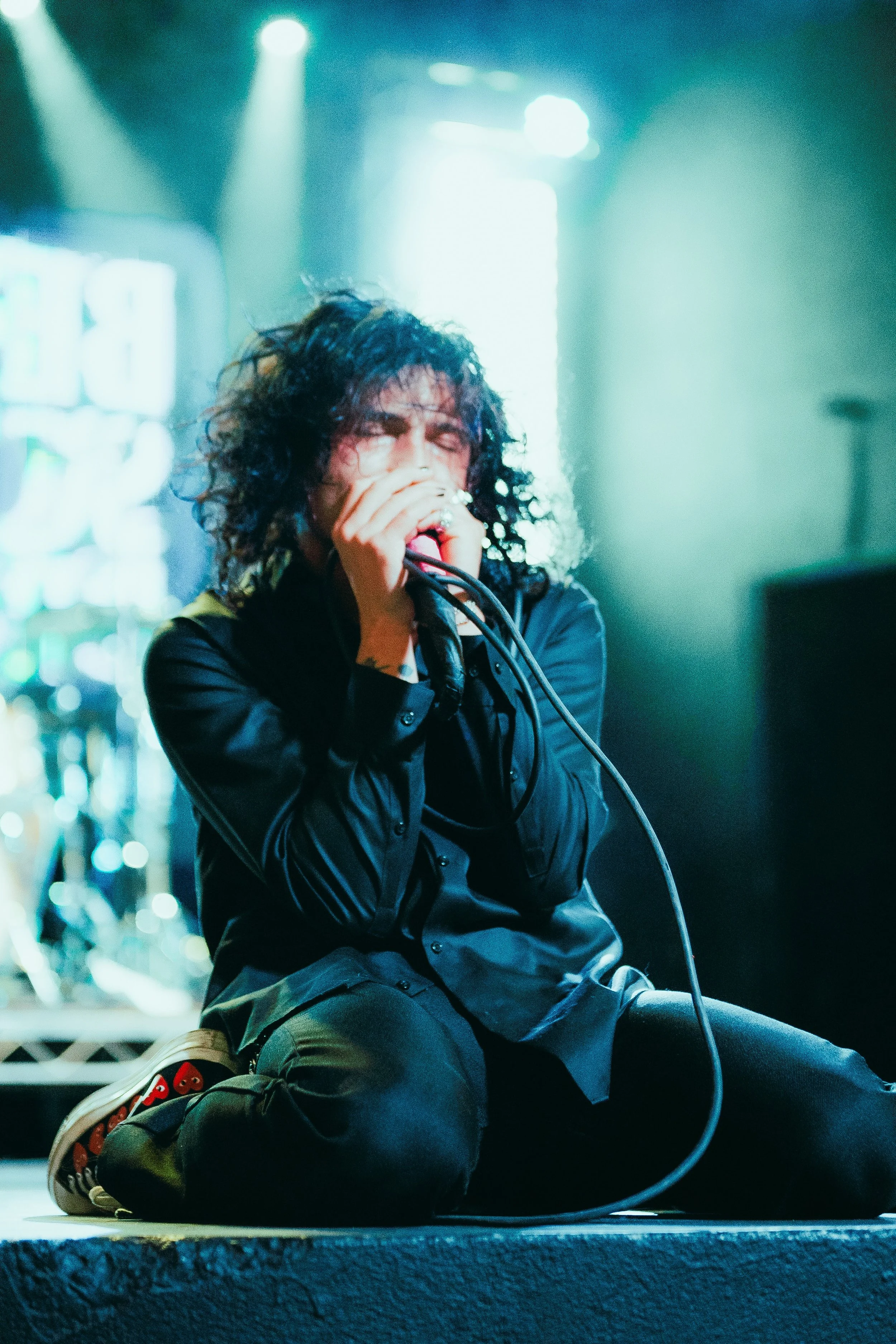 Person with curly black hair singing into a microphone on stage, wearing a black jacket and black pants with red and white sneakers, seated on the stage floor with a blue-toned background and stage lighting.