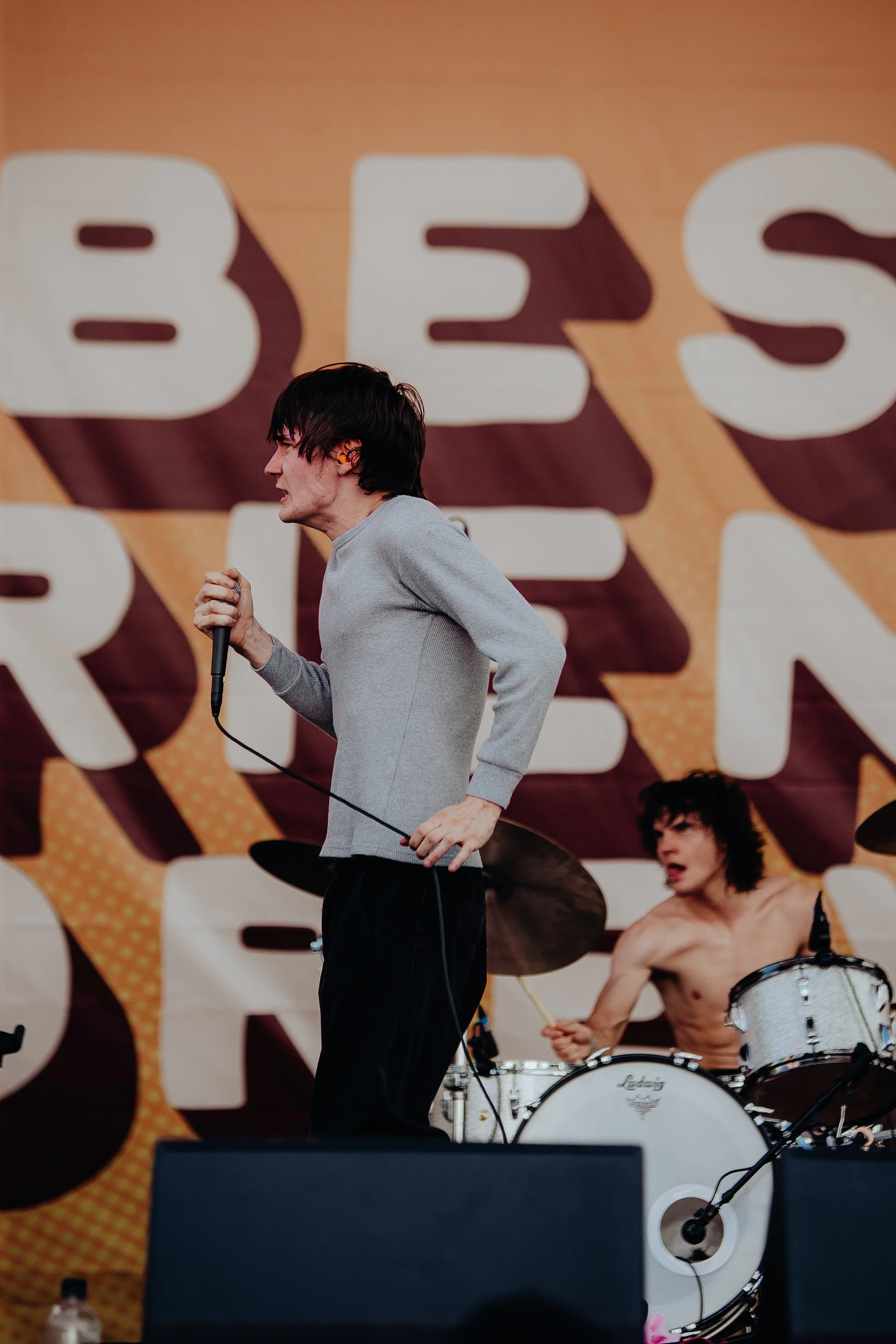A male singer holding a microphone performs on stage with a band. In the background, a drummer is playing drums, and a large sign with bold, colorful letters is visible.