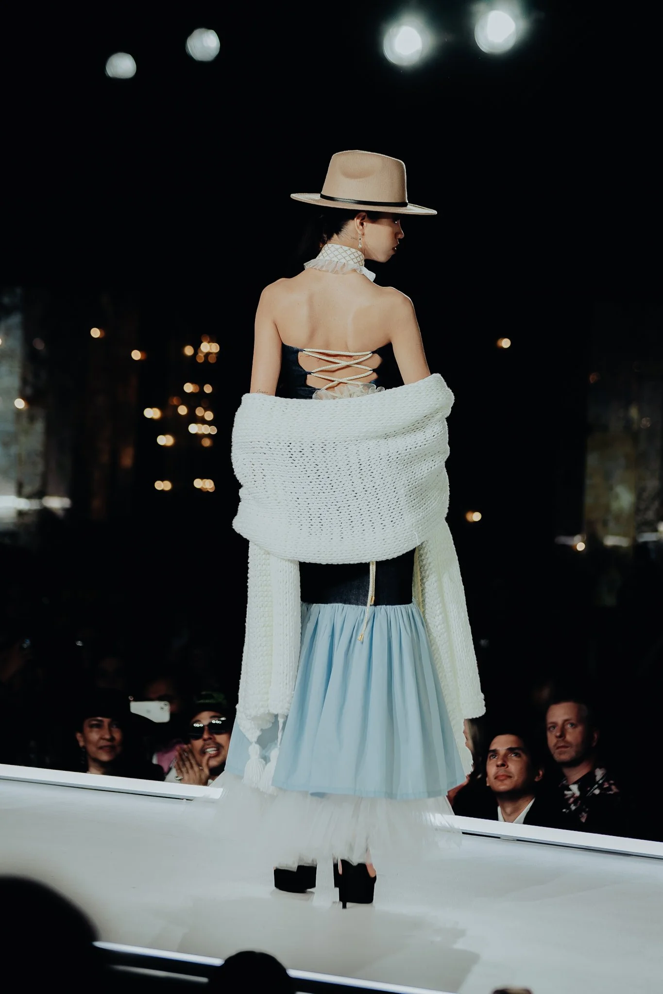 A woman wearing a large beige hat, a black top with lace-up back, and a light blue skirt is standing on a runway at night. She is draped in a white shawl, and spectators are visible in the background, some using their phones.