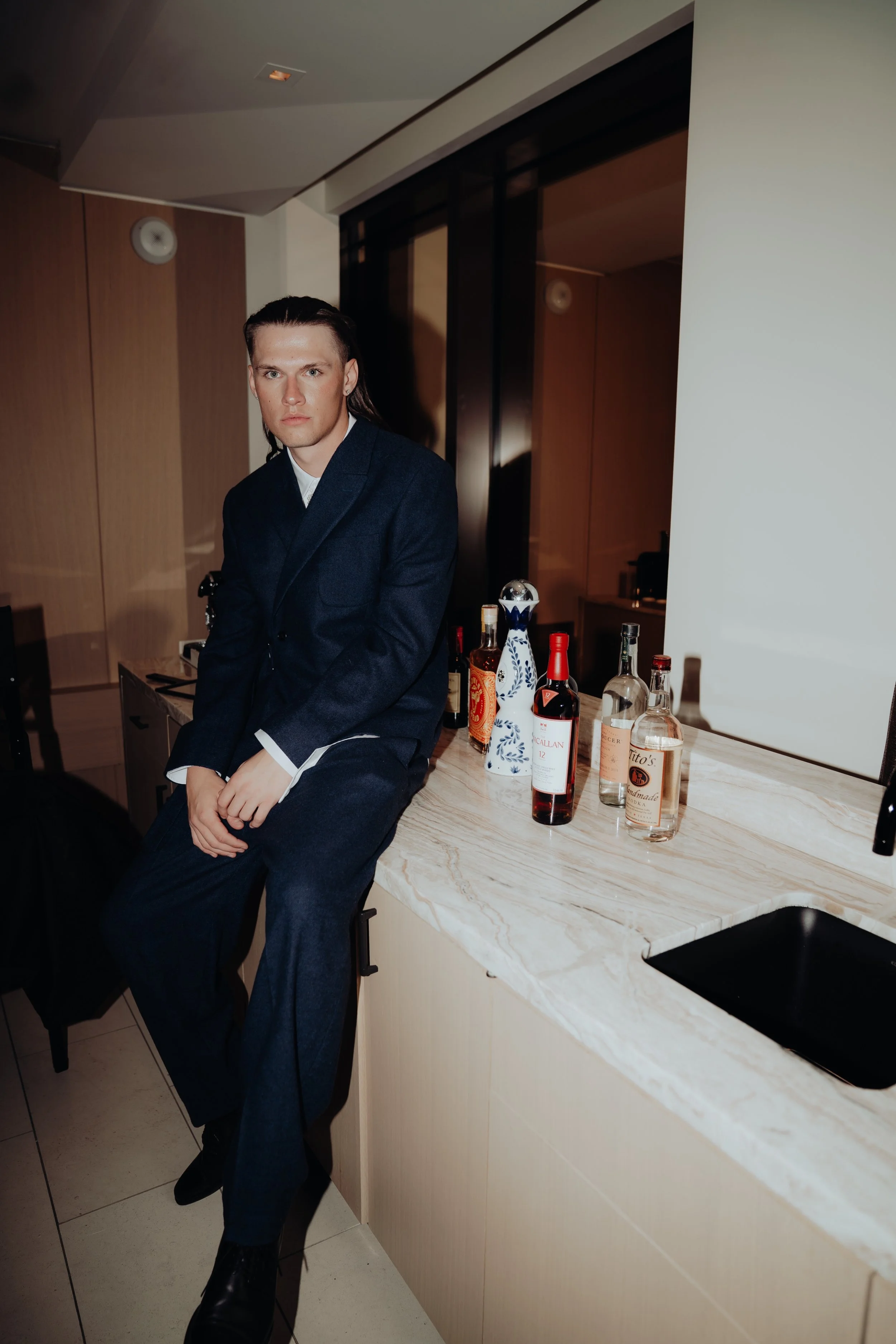 A man with slicked-back hair wearing a dark blue blazer and pants, sitting at a bar counter with various bottles of alcohol on it, in a modern, upscale room with wooden paneling and black accents.