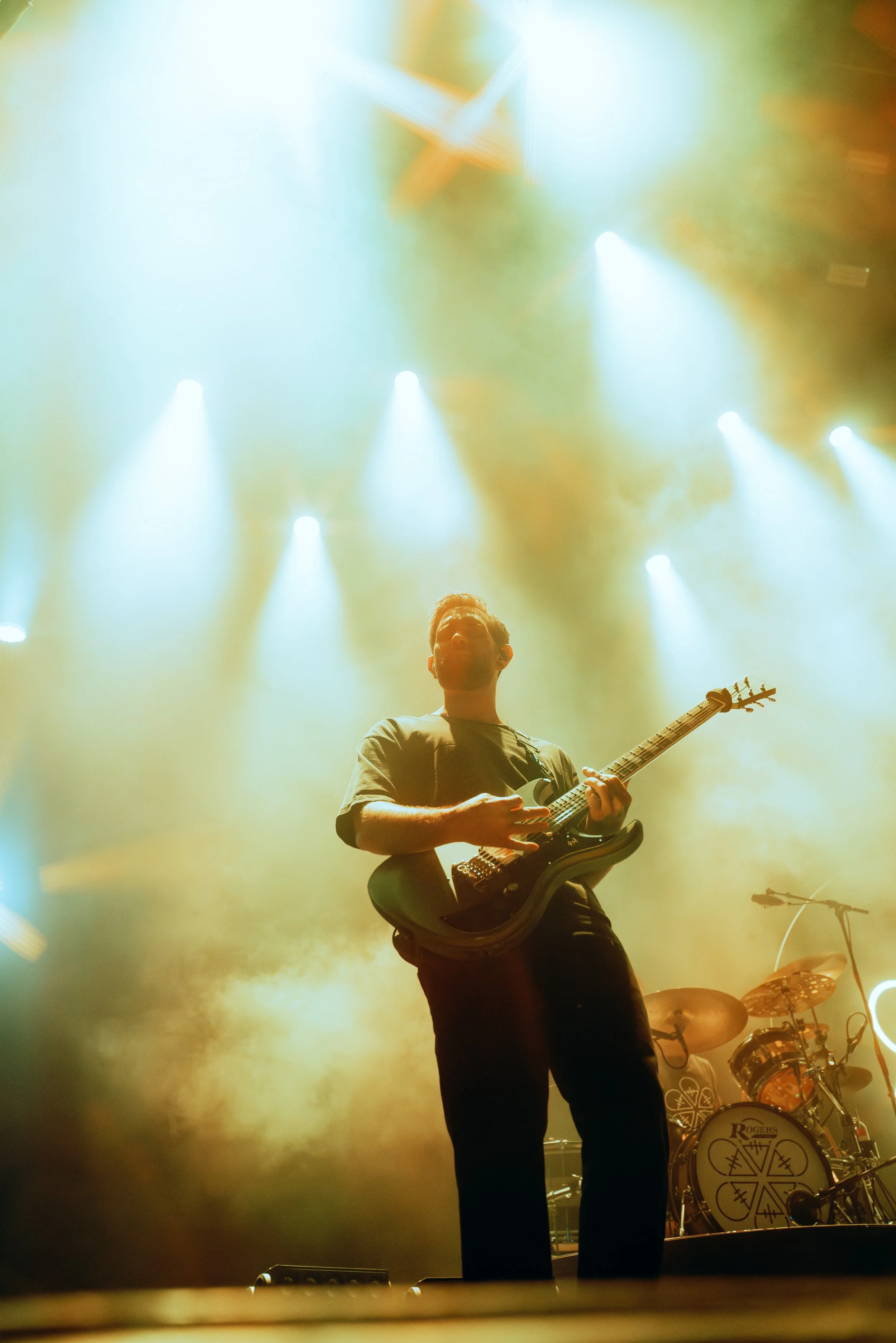 Musician playing an electric guitar on stage with bright stage lights and a drum set in the background.