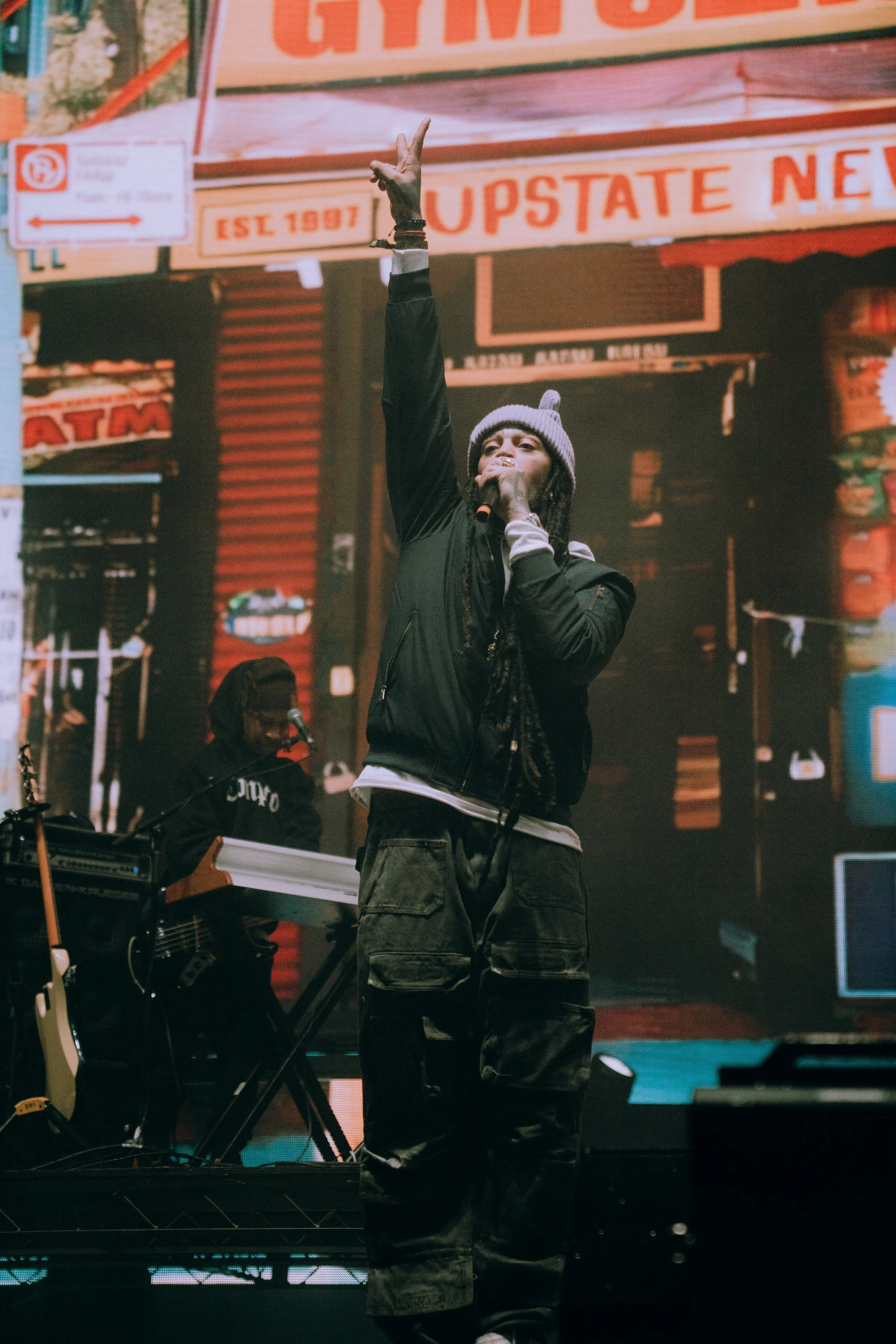 A person performing on stage, wearing a gray beanie, black jacket, and cargo pants, with one arm raised and the other holding a microphone, in front of a city street backdrop.