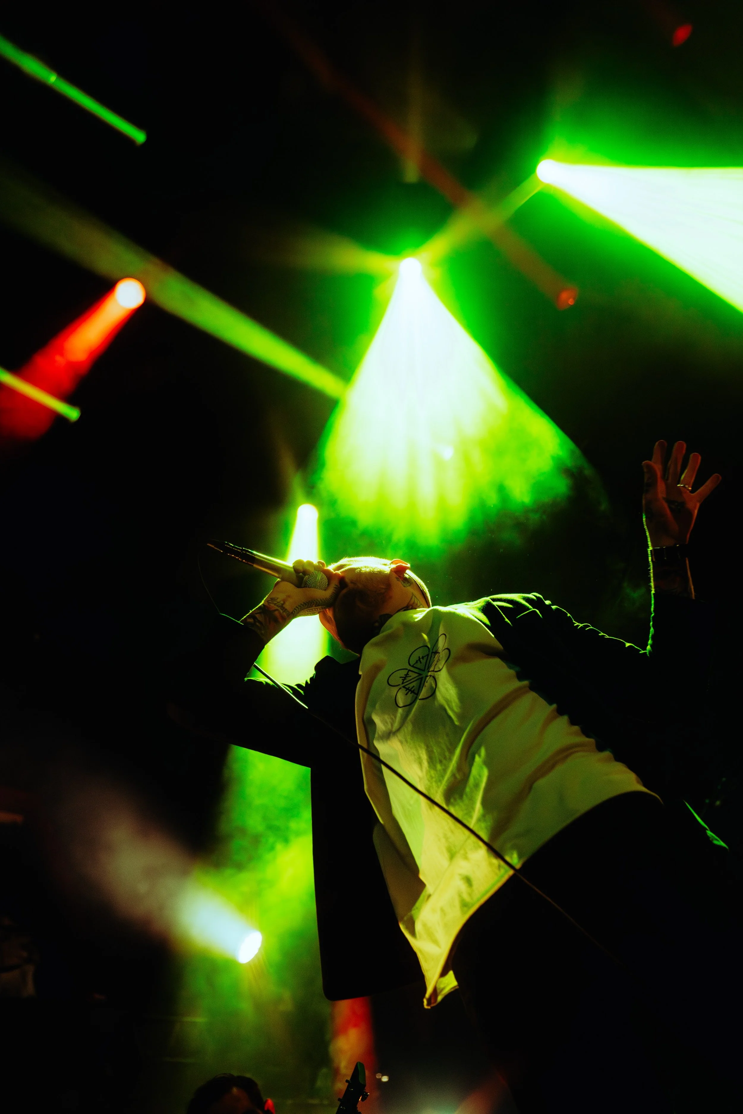 A performer on stage holding a microphone and singing, illuminated by vibrant green and yellow stage lights, with their other hand raised in the air.