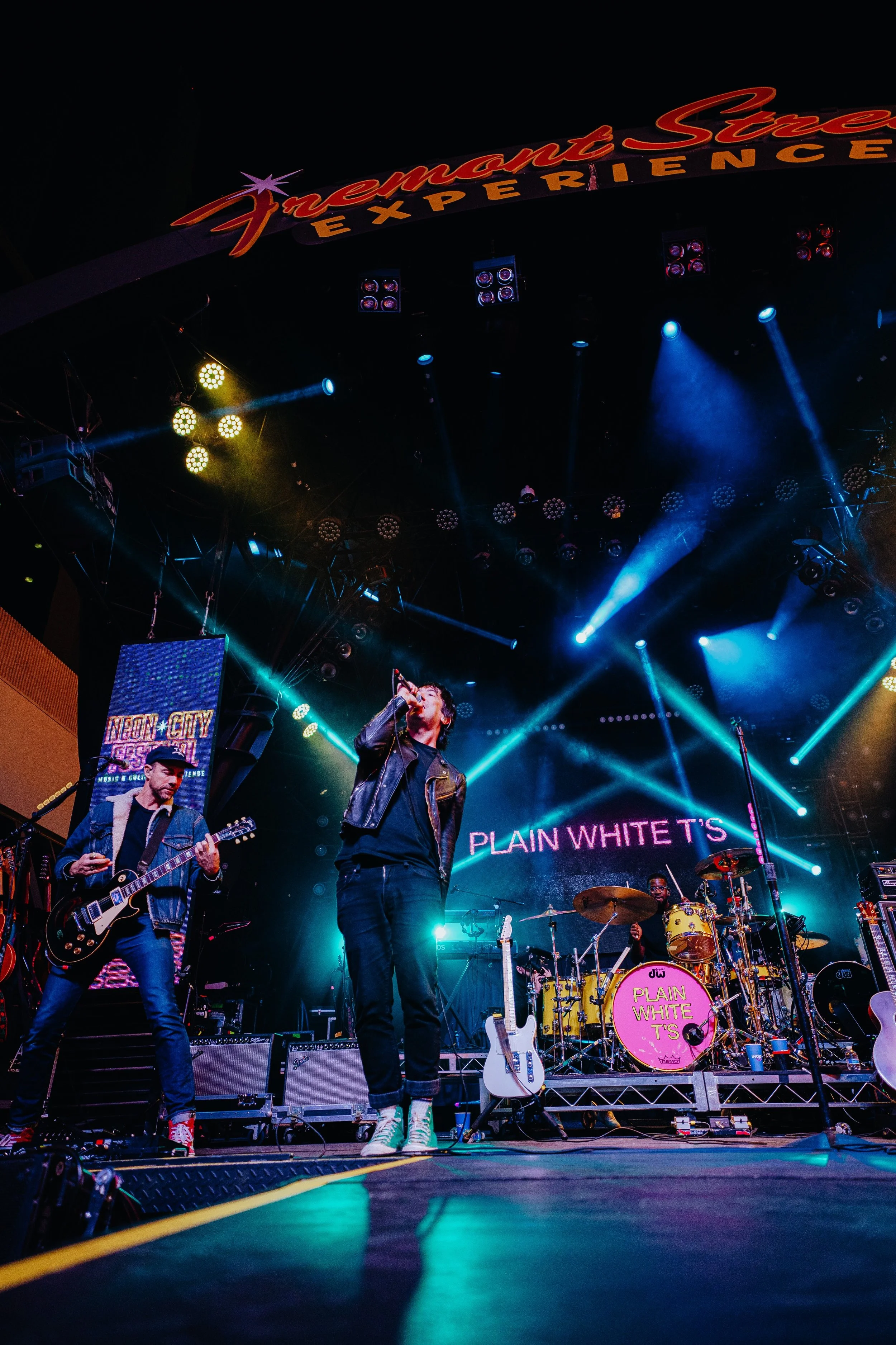 A band performs on stage at a concert at Night Market Street Experience, with a lead singer, guitarist, and drummer, all illuminated by colorful stage lights, and a large sign overhead reading ’Night Market Street Experience’.