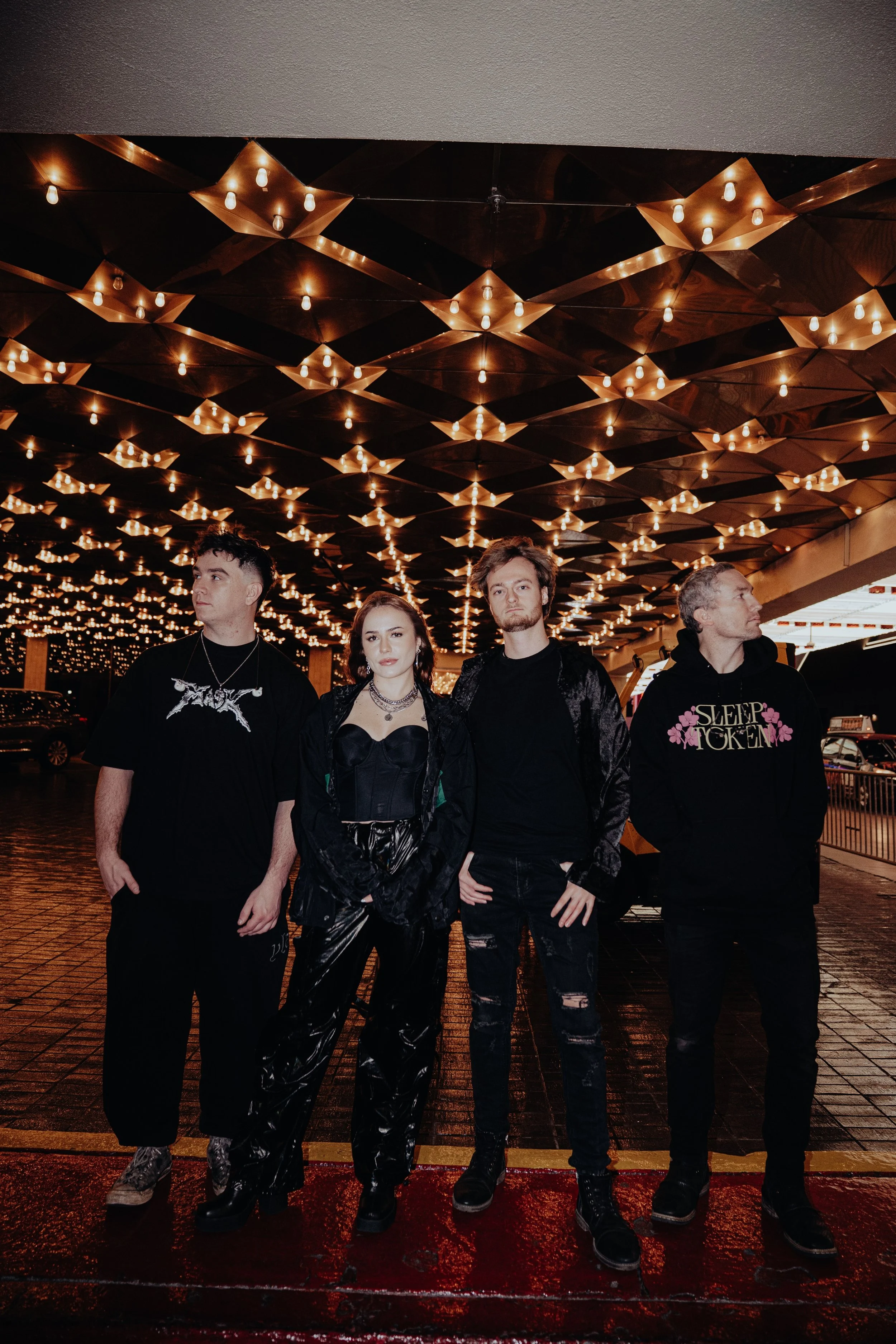 Four young adults standing outside at night under a ceiling decorated with numerous small warm-colored lights. They are dressed in dark, edgy fashion and standing on wet pavement.