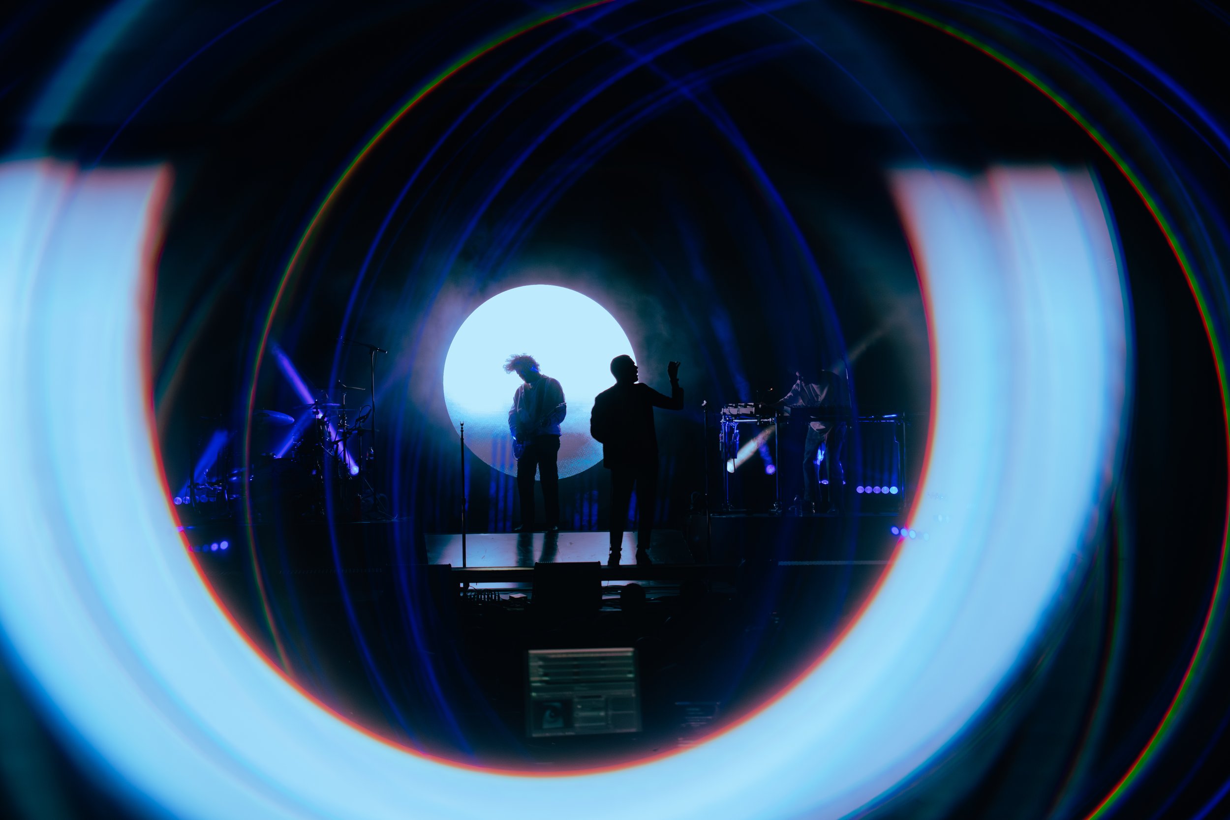 Silhouettes of two people on stage, one with curly hair, microphone, and other with raised hand, with colorful light effects and a large circular display in the background.