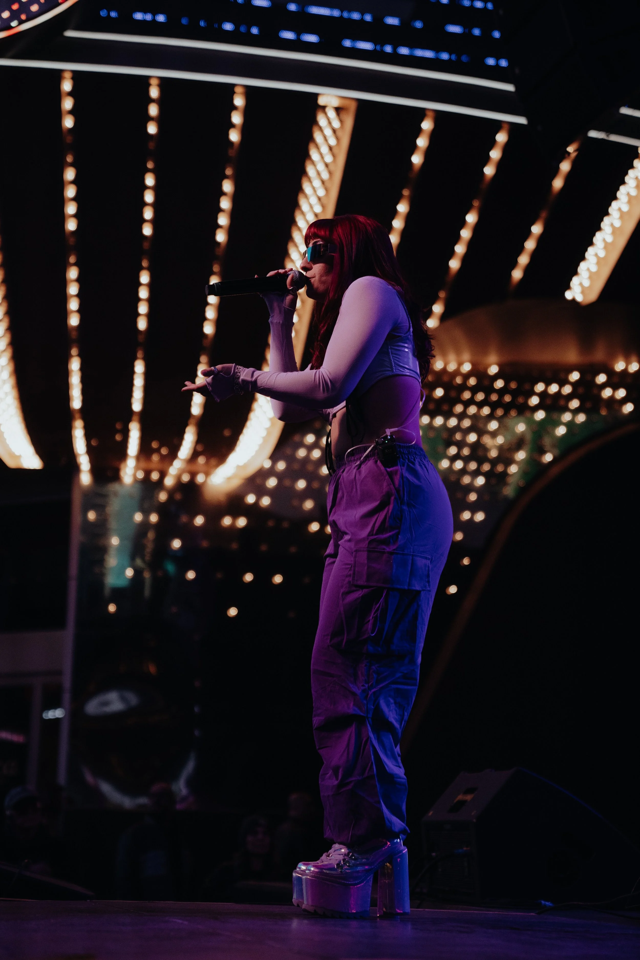 A performer with red hair, wearing sunglasses and purple clothing, stands on a stage at night, holding a microphone, with bright lights in the background.