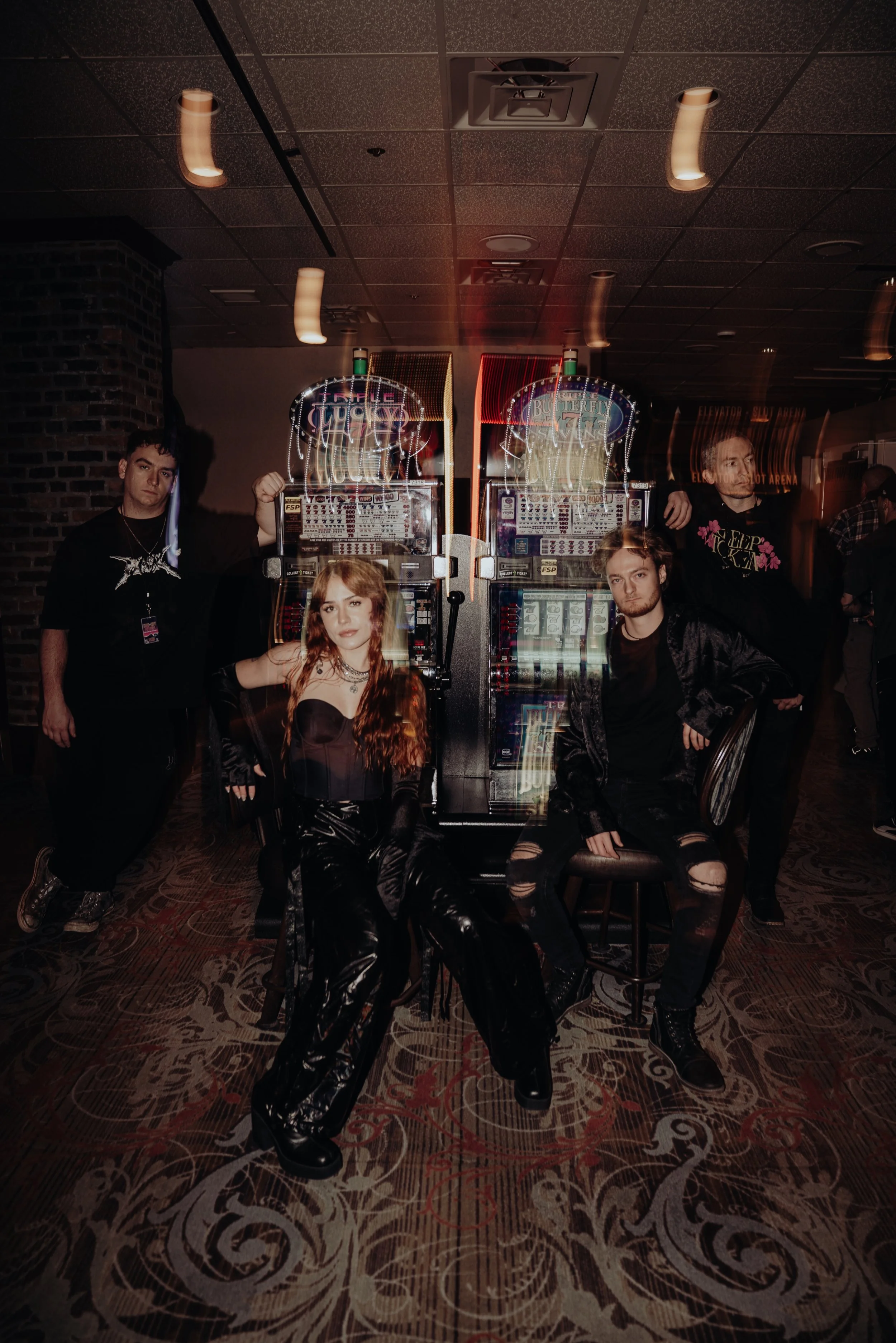 Group of five young adults posing in front of two slot machines in a dimly lit casino or gaming area, with patterned carpet and brick wall visible.