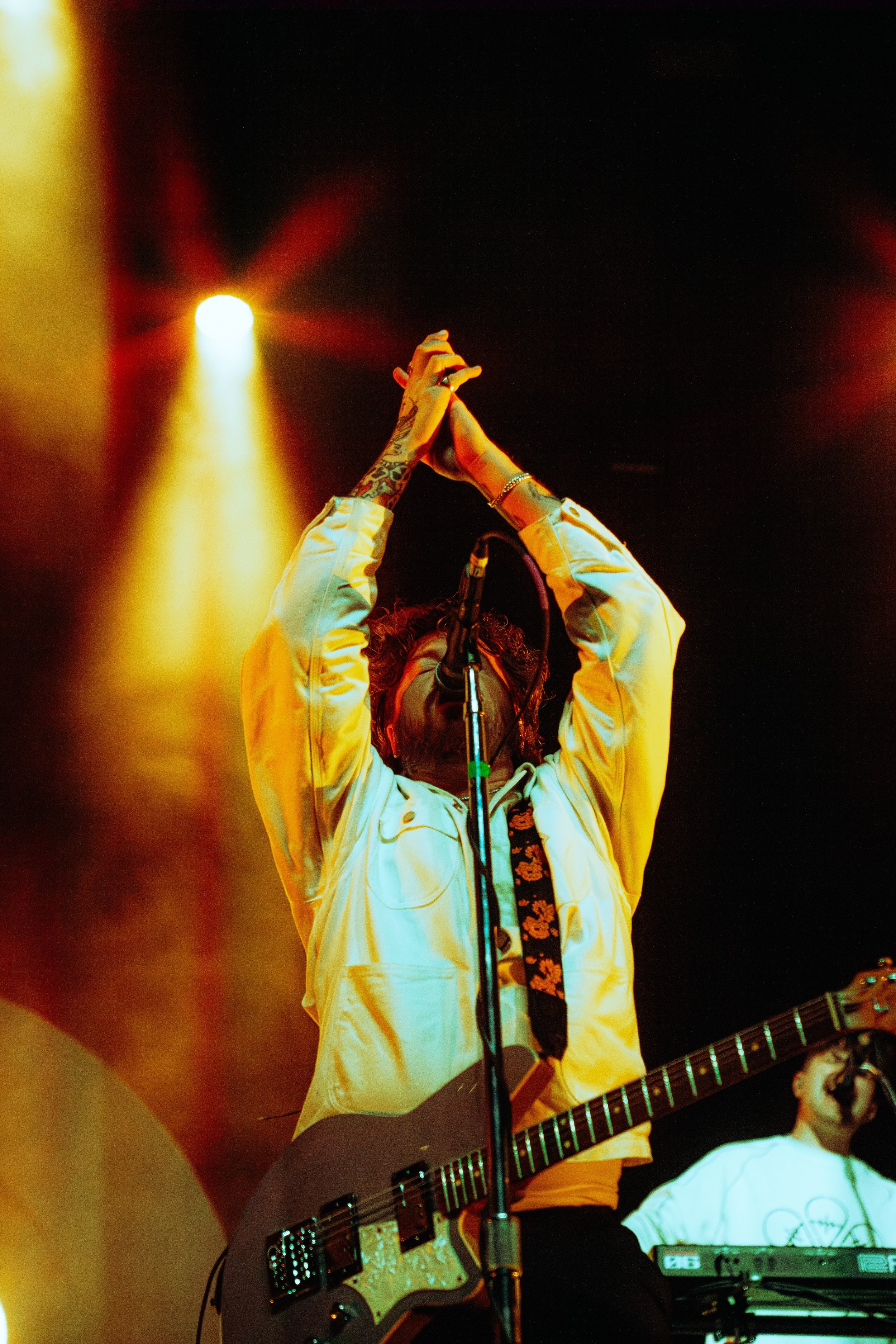A musician with tattoos on their arms, wearing a light-colored jacket, is holding a guitar with a strap and singing into a microphone. There are stage lights behind them, with one bright yellow light on the left. Another person next to them is workin