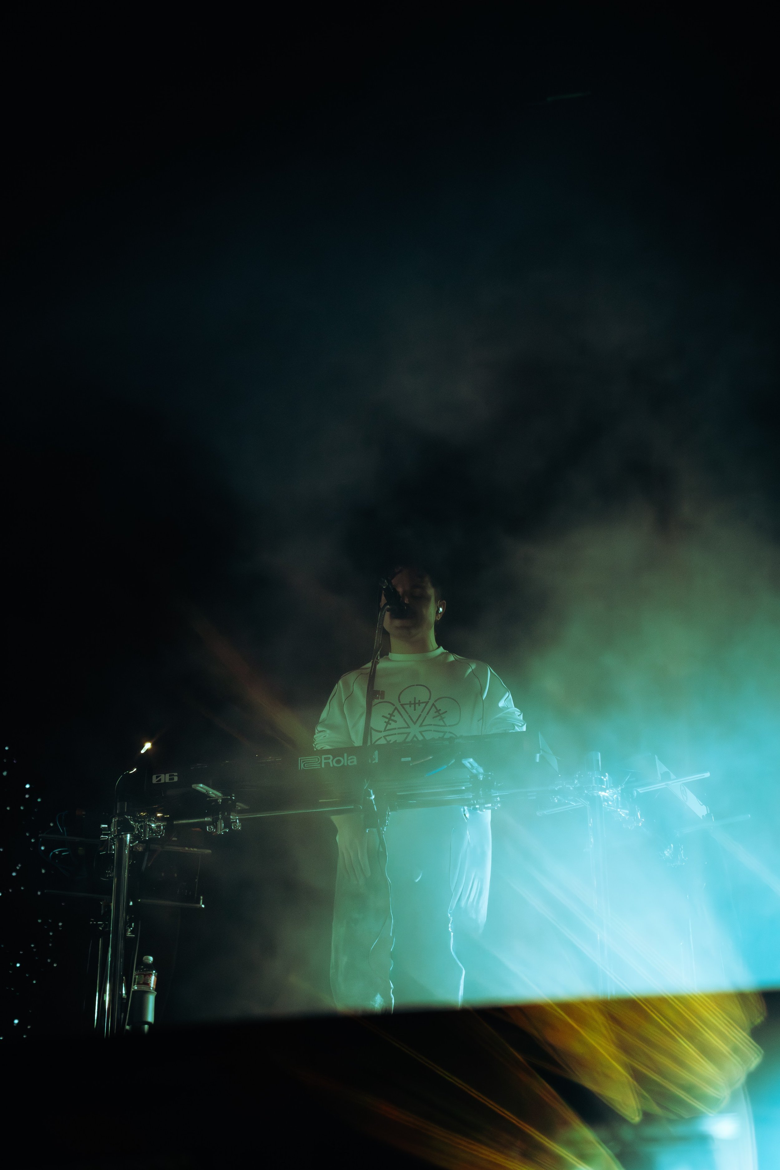 A DJ wearing glasses and a white shirt performing at a nighttime event with green, yellow, and white lights surrounding him, electronic equipment including a Roland keyboard and turntables in front.