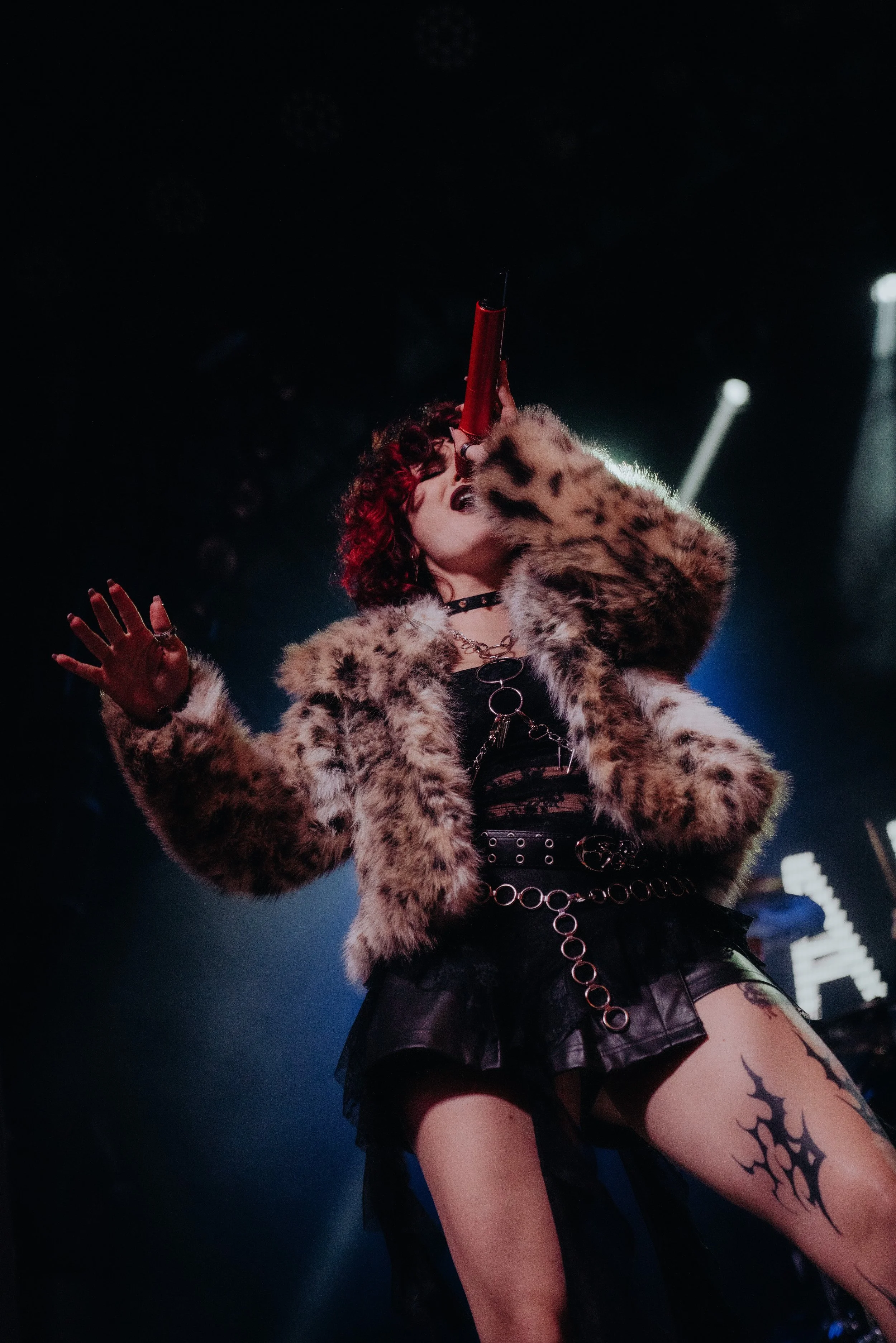 A female performer on stage singing into a microphone, wearing a fur coat, a choker, a leather top, a leather skirt, and playing with body tattoos.