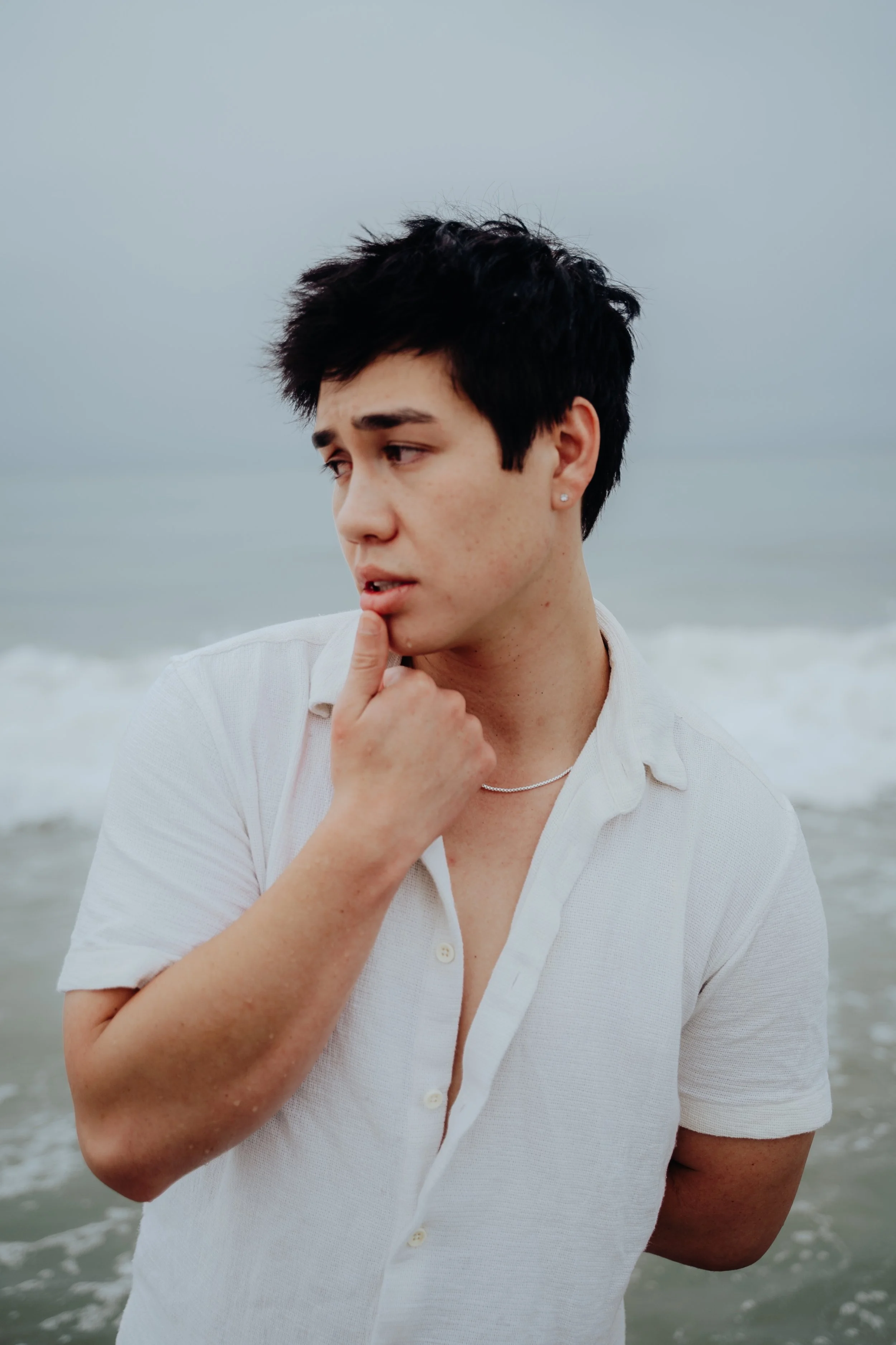 A young man with short dark hair, wearing a white button-up shirt, standing on the beach with the ocean and cloudy sky in the background. He is holding his finger to his lips, appearing thoughtful or pensive.
